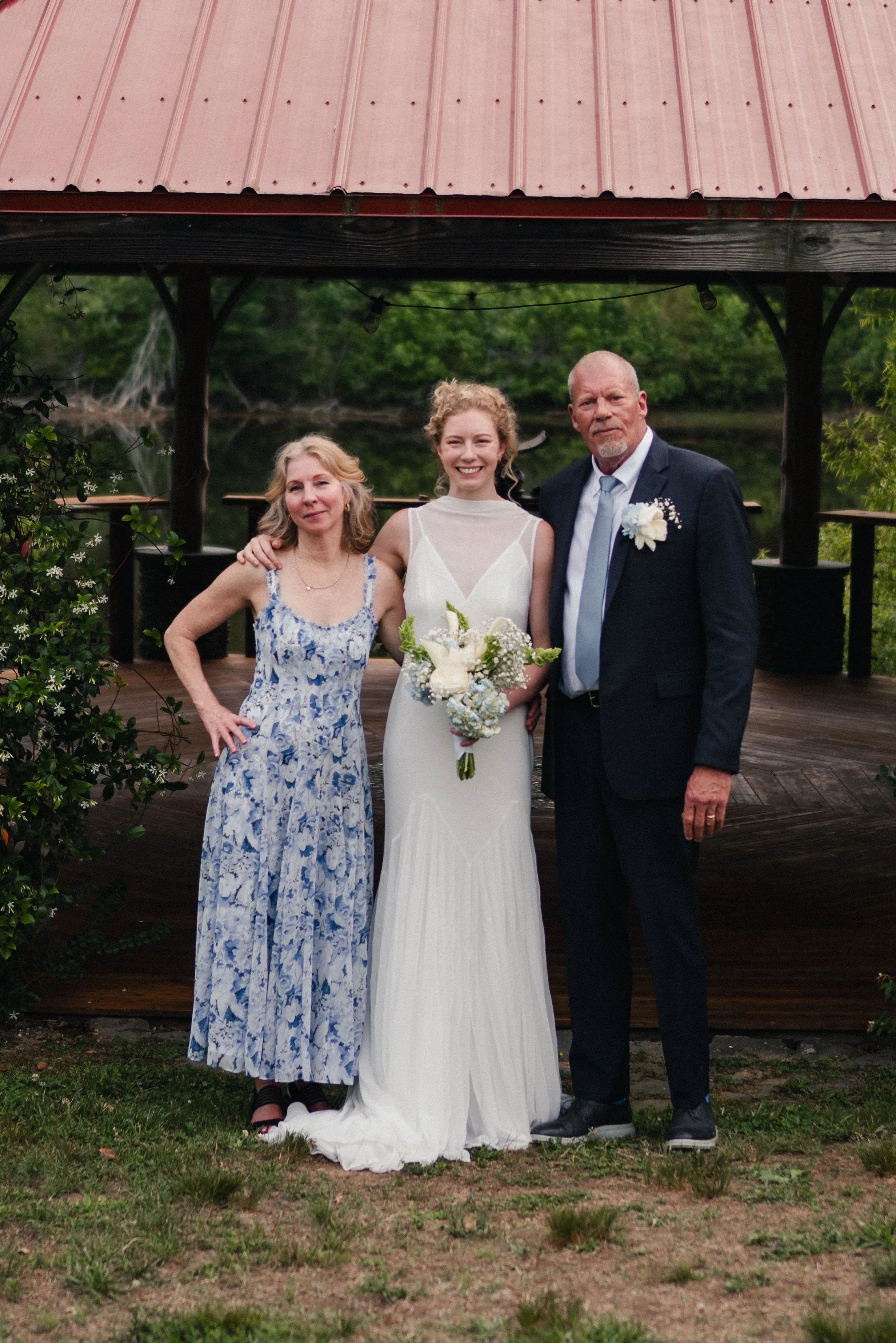 Three people standing outdoors on a grassy area in front of a wooden structure with a red metal roof. Two women and one man, all dressed in formal attire, smiling for a photo. The woman in the center is wearing a white dress and holding a bouquet of 