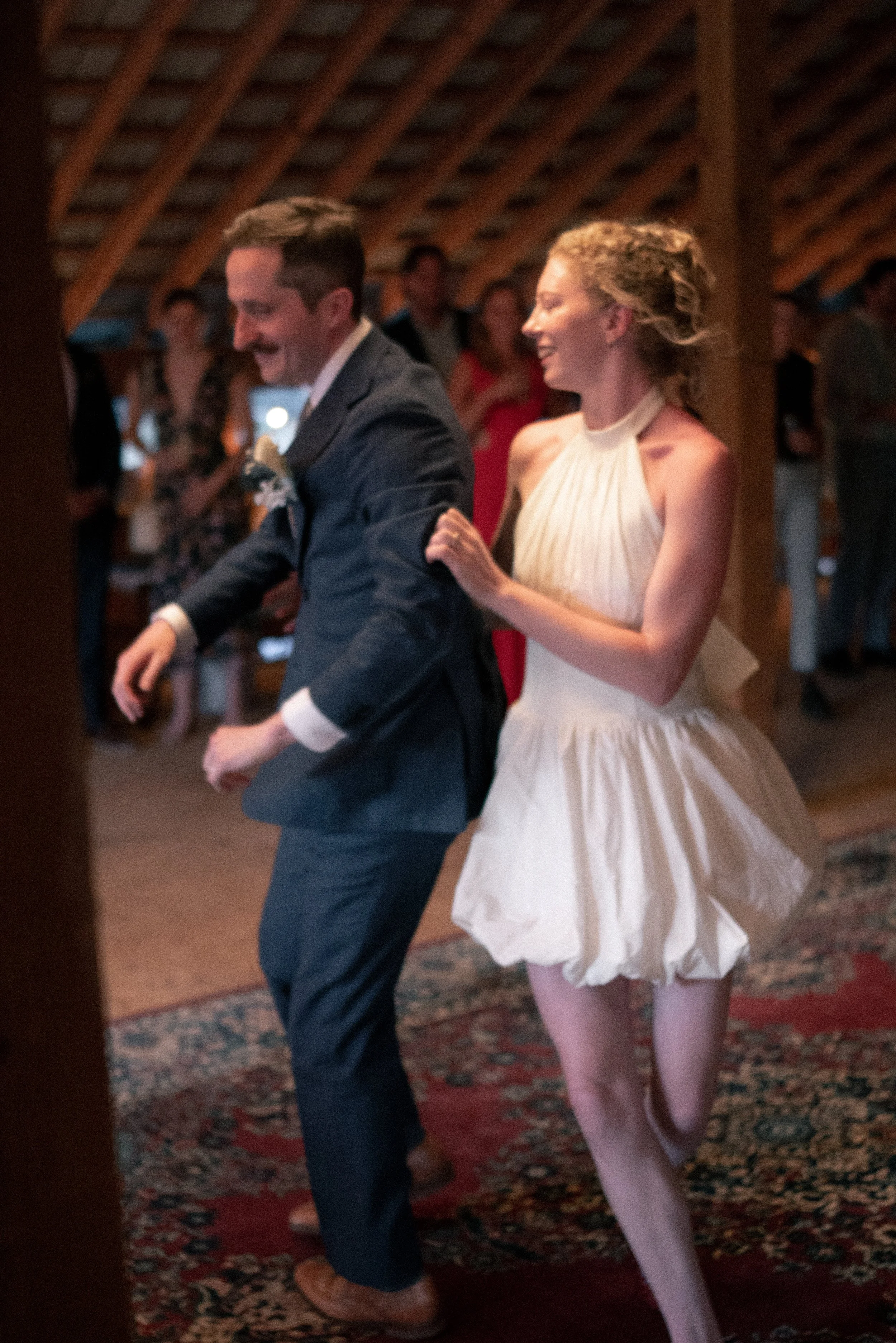 A couple dancing at a wedding reception, with the woman wearing a sleeveless white dress and the man in a dark suit. They are smiling and holding hands, with guests watching in the background.
