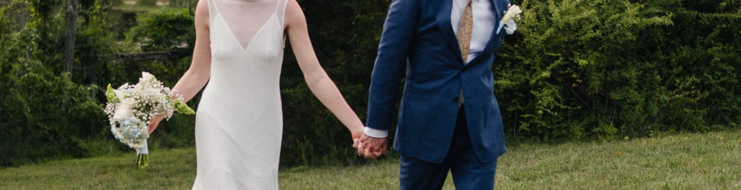 A bride and groom holding hands outside during their wedding ceremony, with greenery in the background.