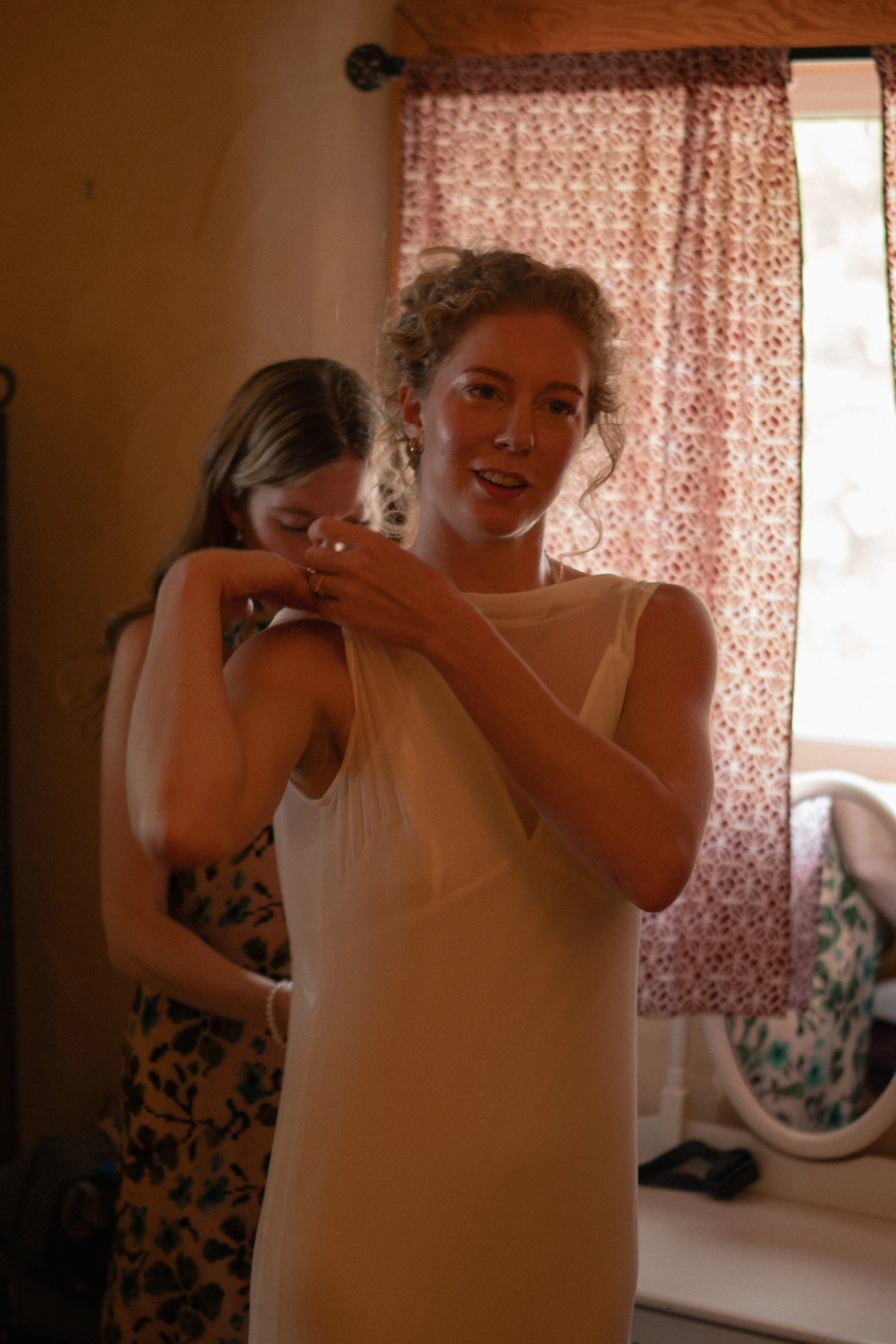 A woman wearing a light-colored dress adjusting her clothing, with another woman in the background, in a room with curtains and a laundry basket.