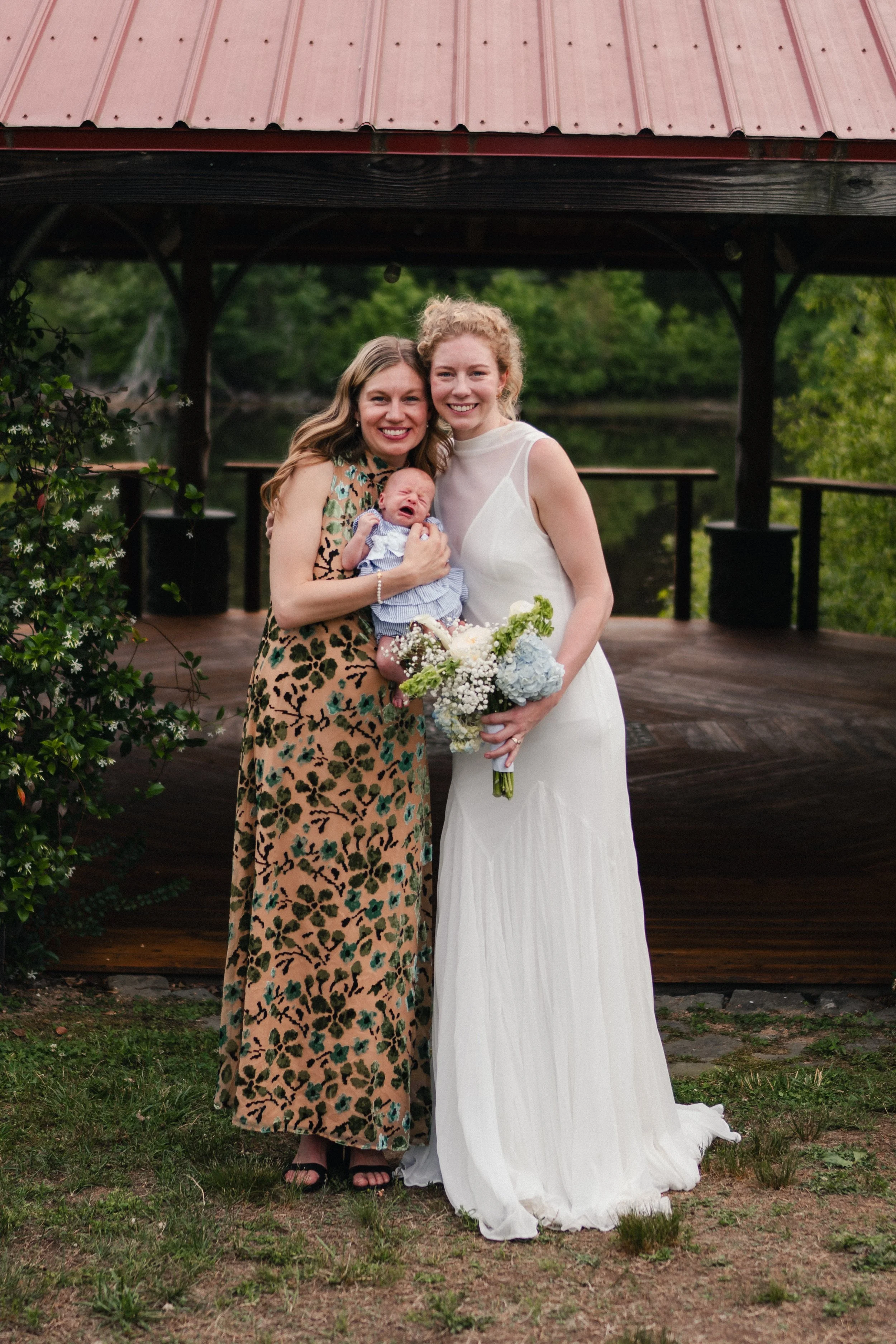 Two women, one holding a crying baby, smiling and standing on a grassy area near a wooden gazebo, with greenery in the background, at what appears to be a wedding or special event.