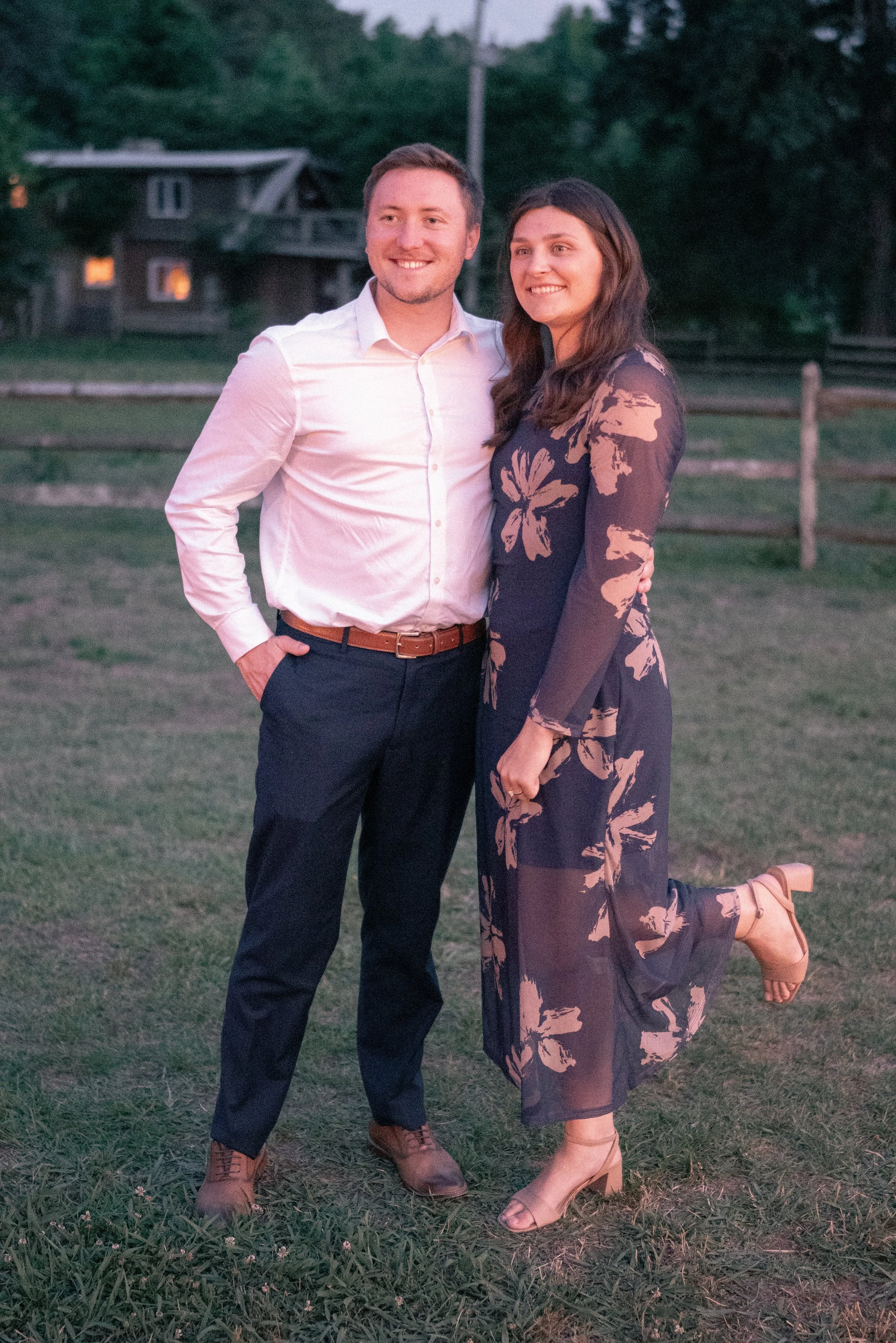 A happy couple standing on grass with greenery and a house in the background at dusk. The man is wearing a white dress shirt and dark pants, and the woman is wearing a dark floral dress and heels. She is kneeling on one leg with a smile, and the man 