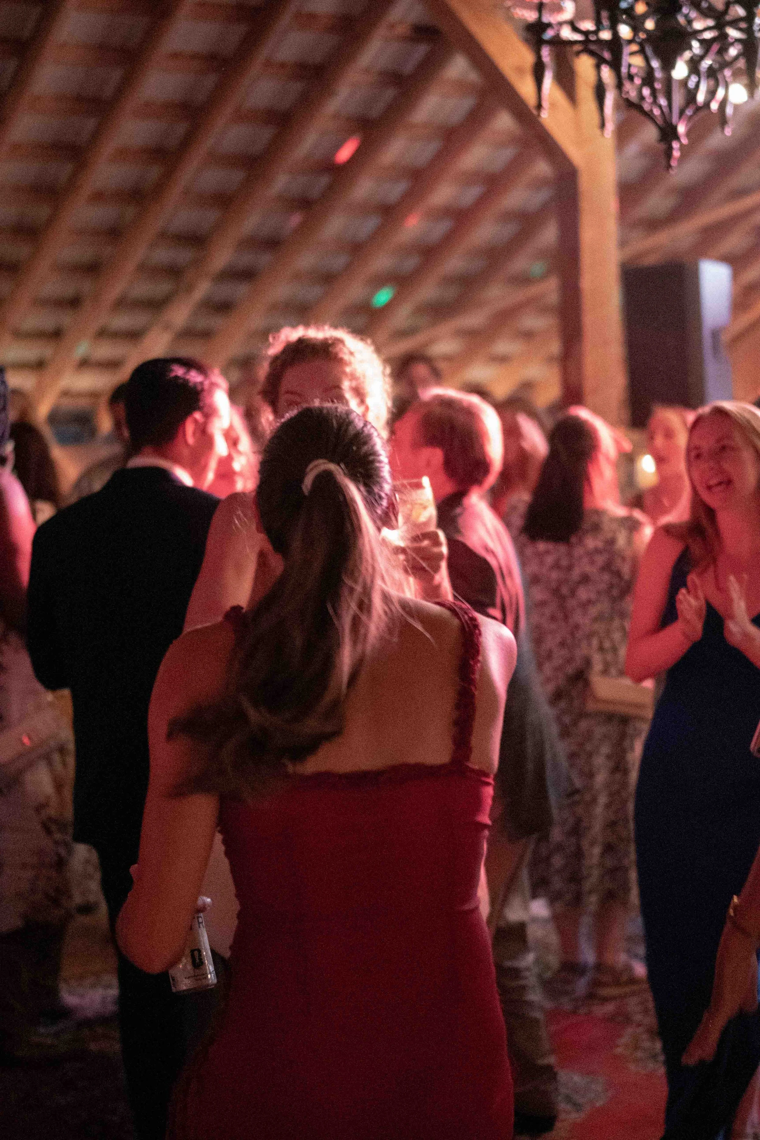 People at a party or wedding reception, dressed in formal attire, socializing in a warmly lit venue with exposed wooden beams and a chandelier.