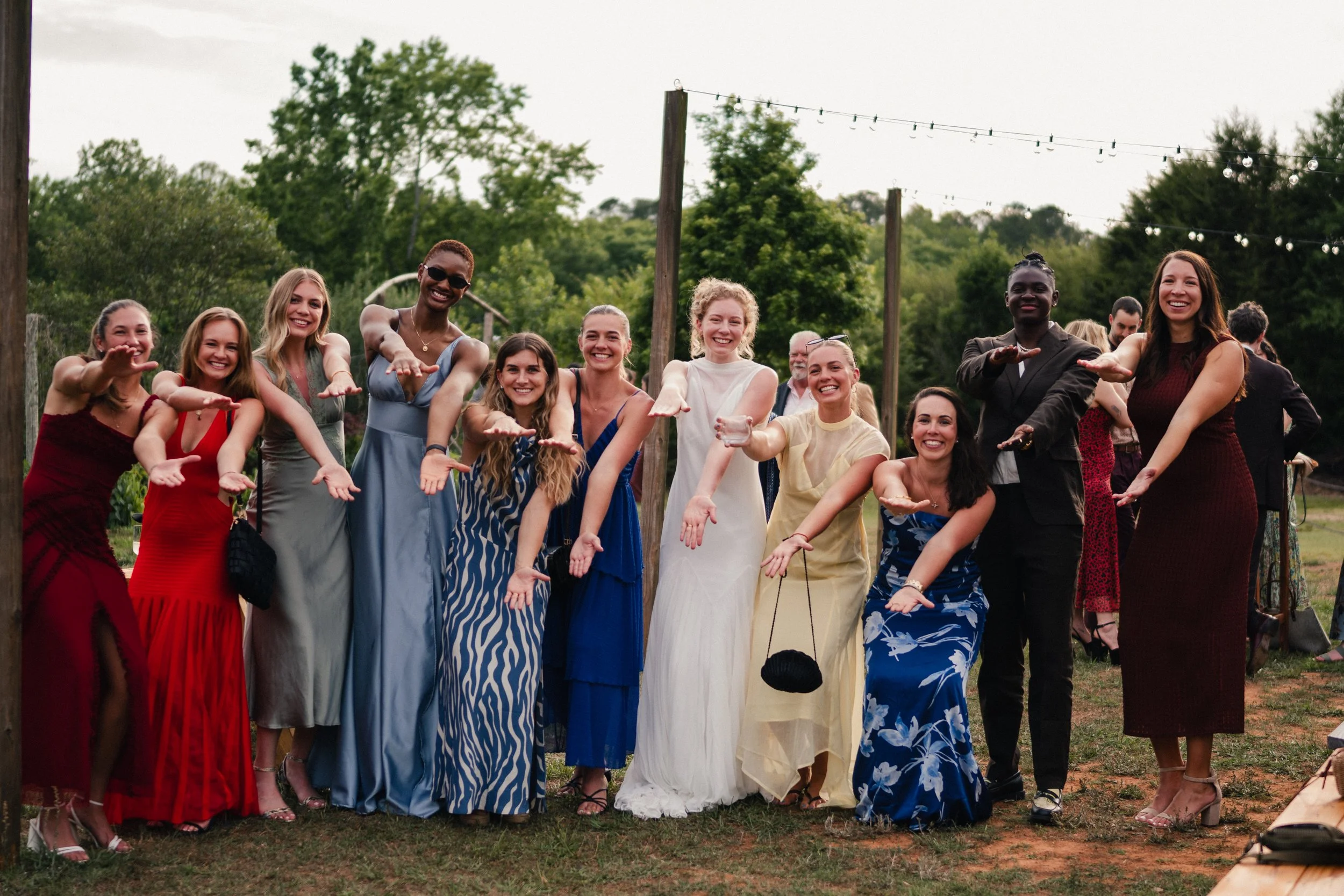 A group of women and men at an outdoor gathering, smiling and extending their arms forward, with a background of trees, string lights, and a cloudy sky.