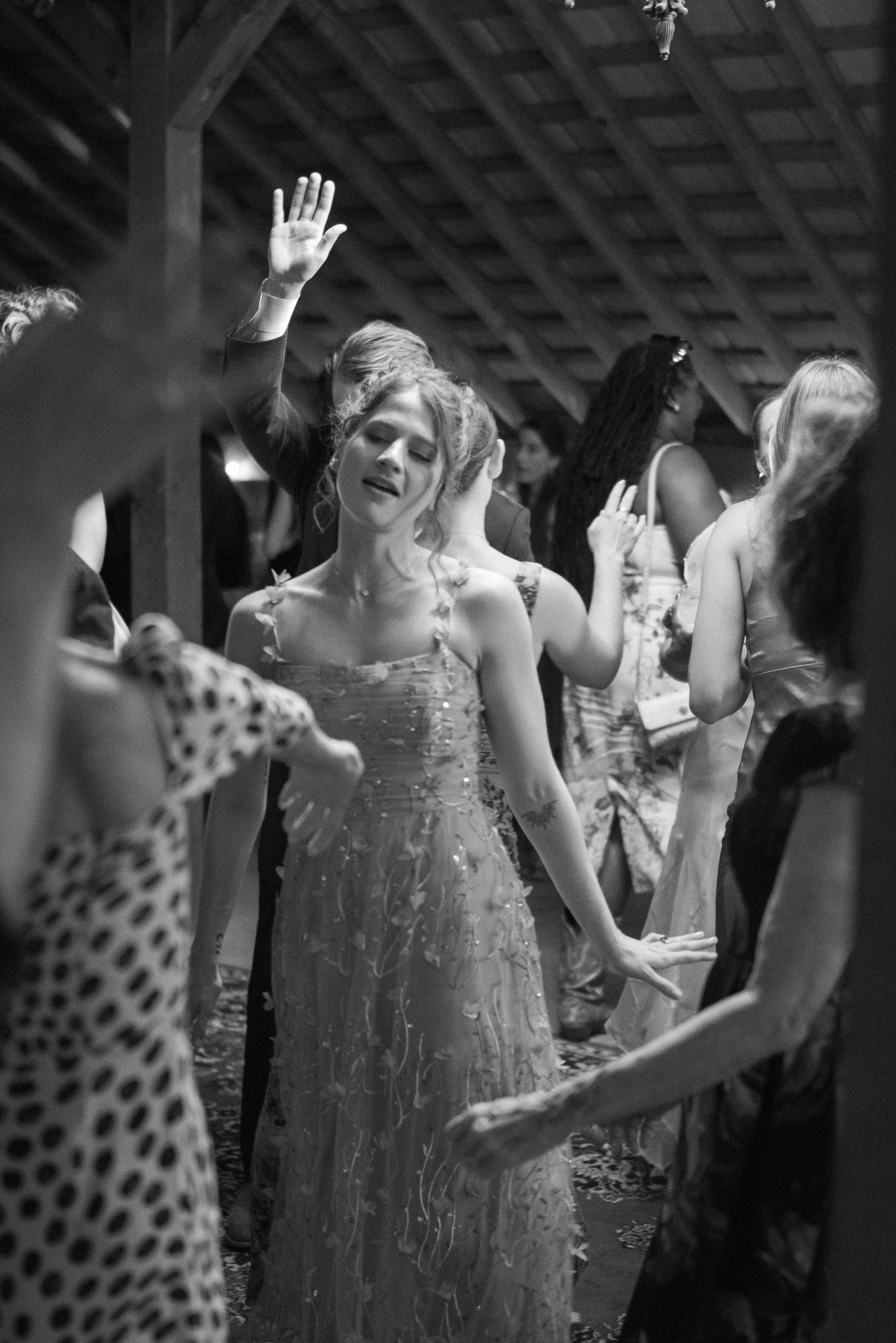 Women dancing at a party or reception, with some women raising their hands and closing their eyes. The setting appears to be indoors with a wooden vaulted ceiling.