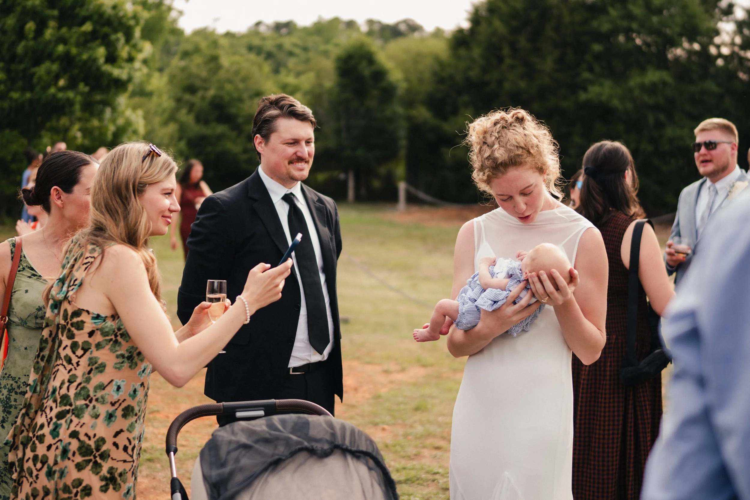 A group of people at an outdoor event, with women and men chatting, and a woman holding a baby, dressed in formal and casual attire, in a green park setting.