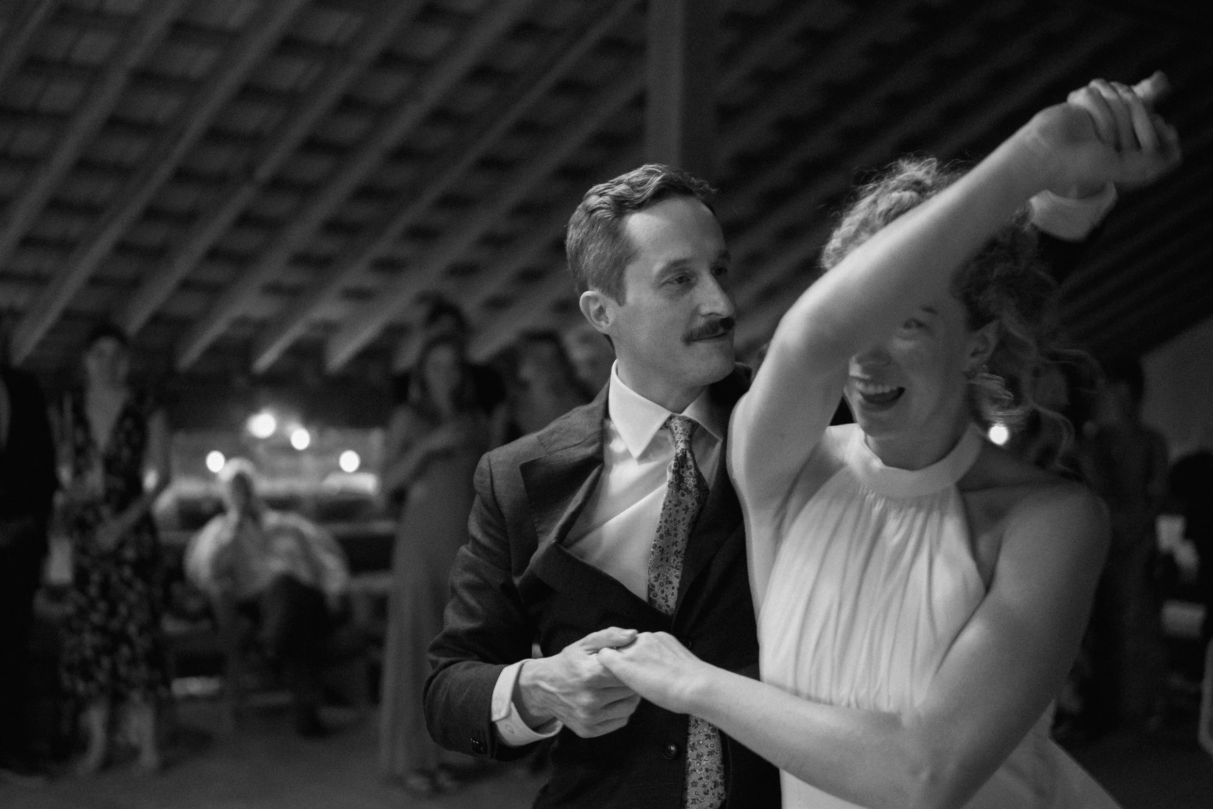 A black and white photo of a man and woman dancing closely at a party or dance hall, with other people and a rustic ceiling in the background.