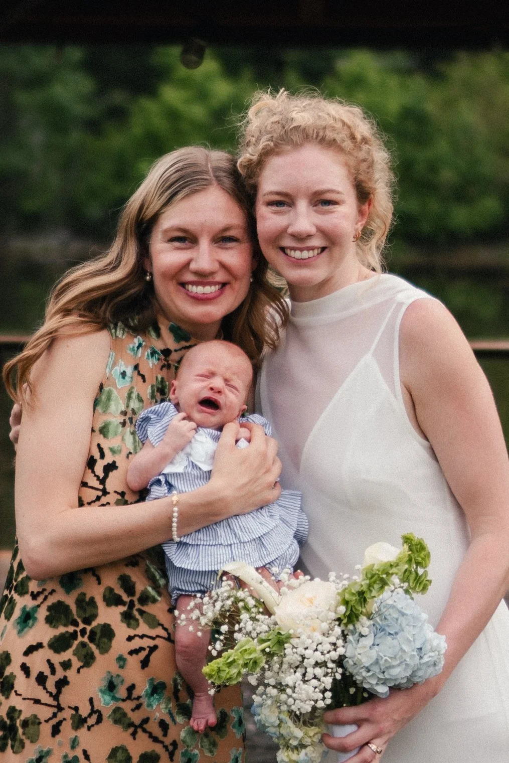Two women smiling and a crying baby, holding a bouquet of flowers outdoors in front of a green background.