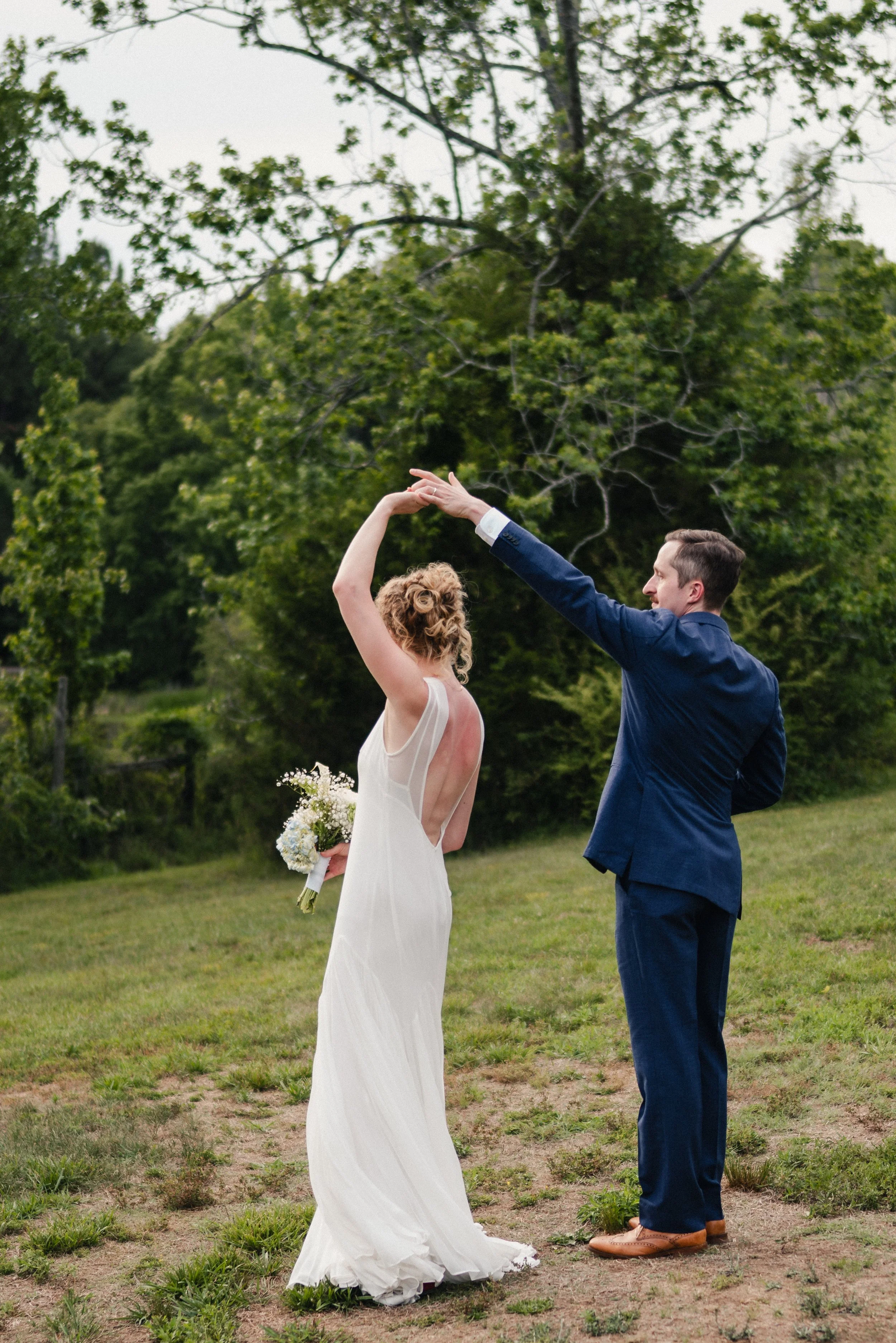 A wedding couple dancing outdoors during daytime, with a lush green background of trees and grass. The bride is wearing a white wedding gown and holding a bouquet of flowers, while the groom is dressed in a blue suit and brown shoes.