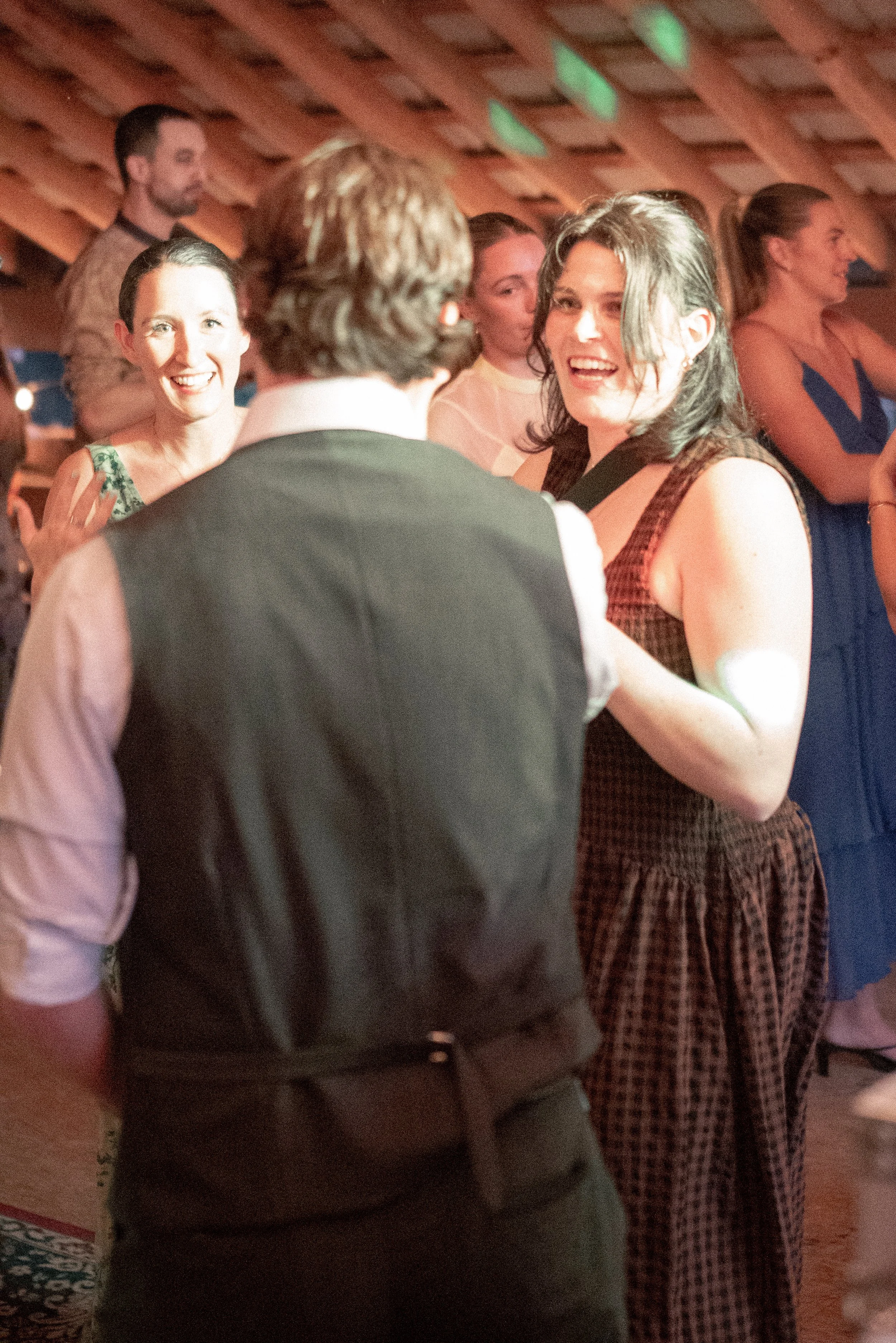 Group of people socializing at an indoor gathering, mostly women, with a woman in a checkered dress smiling and talking to a man in a vest and shirt.