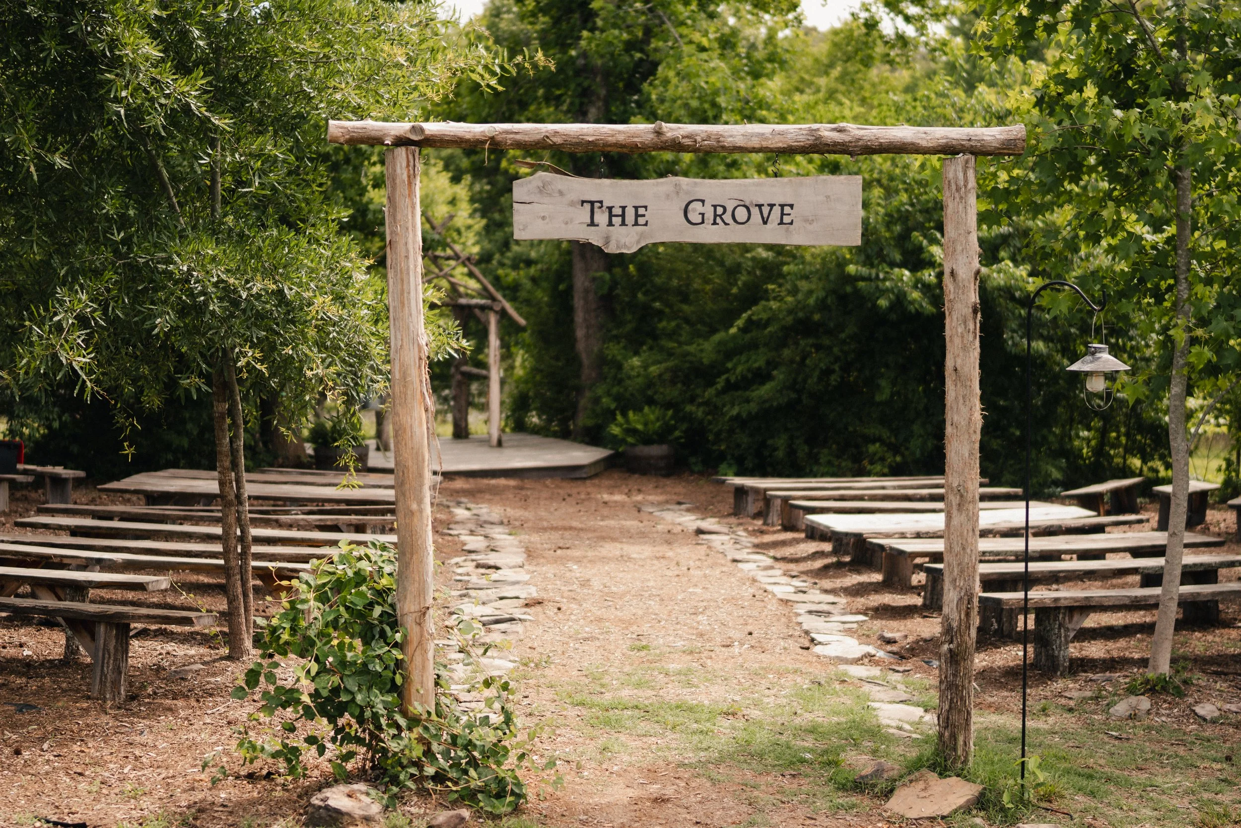 Wooden sign reading 'The Grove' hanging above a dirt path surrounded by green trees and outdoor seating on either side.