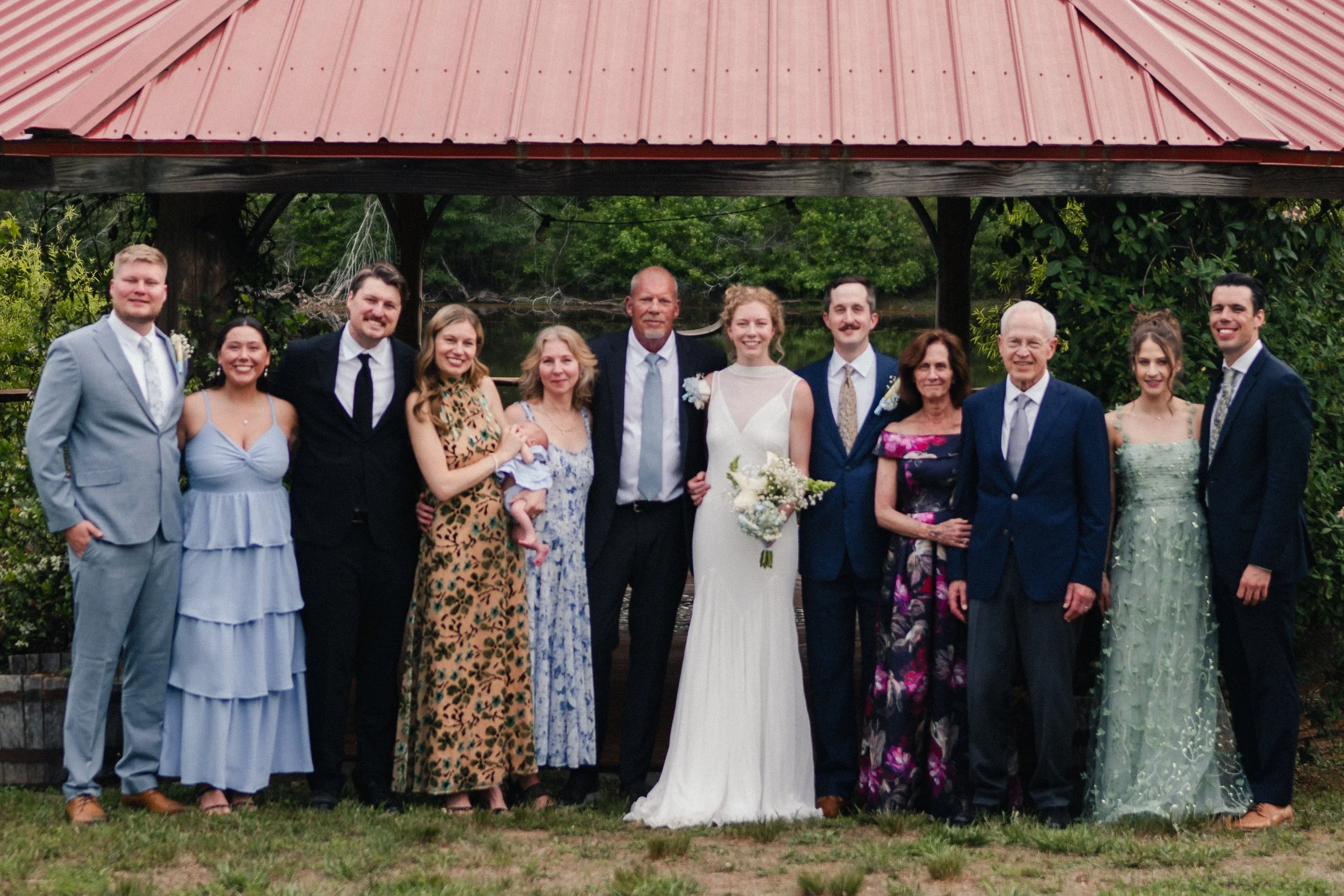 Group of people dressed in formal attire standing outdoors under a red metal roof gazebo with greenery in the background, likely celebrating a wedding.