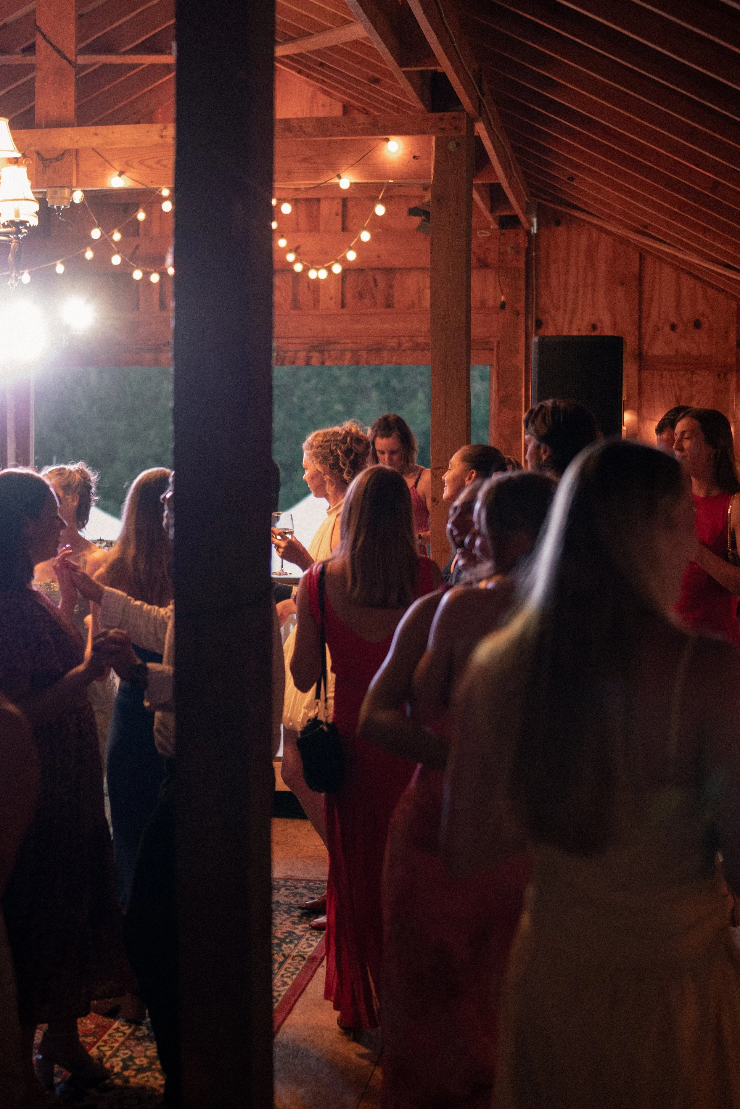 A group of people at a gathering or party inside a wooden structure with string lights hanging from the ceiling. Some individuals are holding drinks and engaging in conversation.