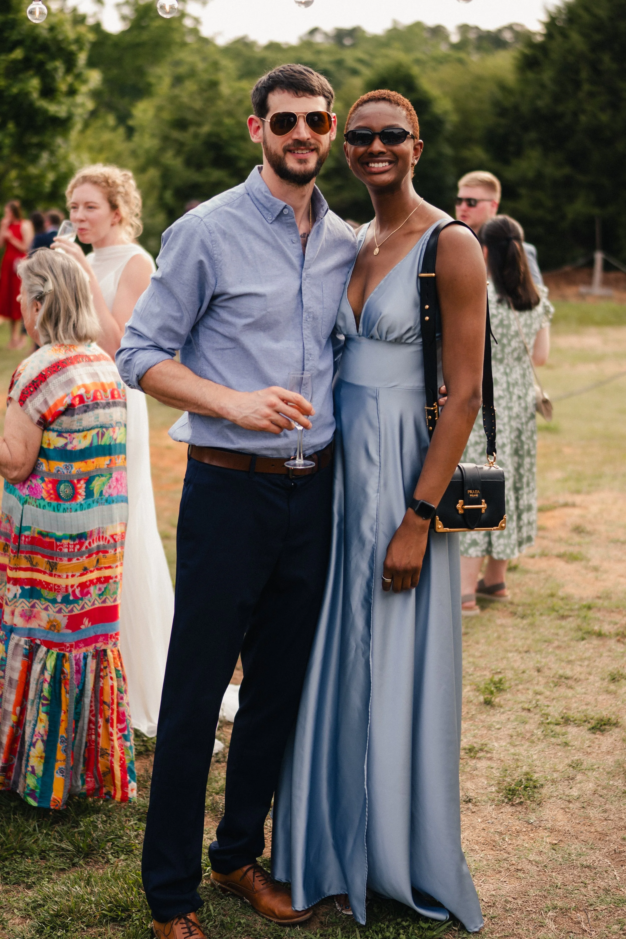 A man and woman smiling at an outdoor gathering, with the man holding a glass of champagne. The man has short dark hair, a beard, sunglasses, a light blue button-up shirt, dark pants, and brown shoes. The woman has short reddish hair, glasses, a gold