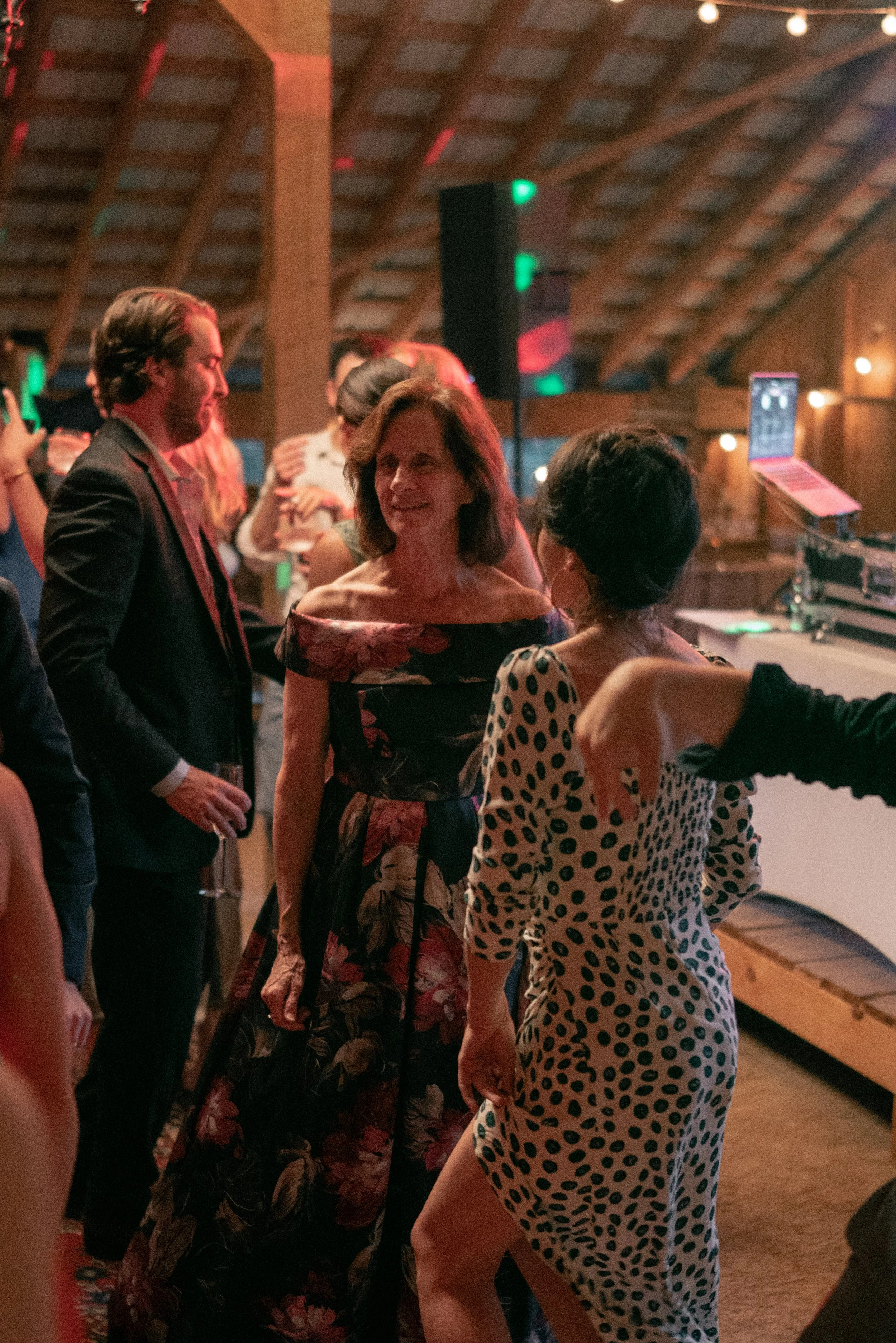People socializing at a party or event in a wooden venue, dressed in formal and semi-formal attire, with a DJ booth and colorful lighting in the background.