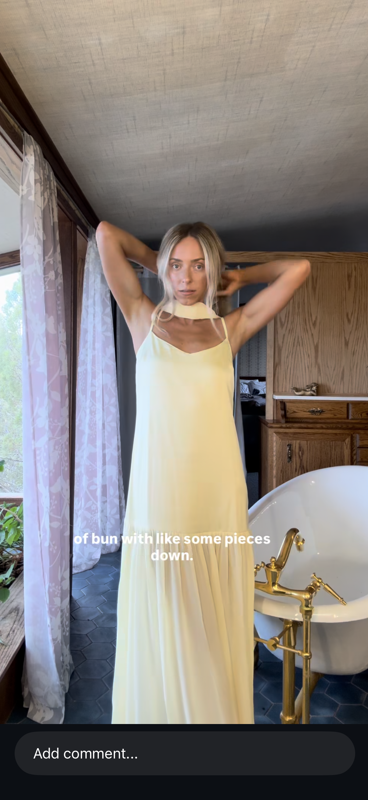 A woman in a yellow dress standing in a bathroom, adjusting her hair, with a bathtub and wooden cabinets in the background, and curtains on a window to the side. Text on the image reads: "of bun with like some pieces down."