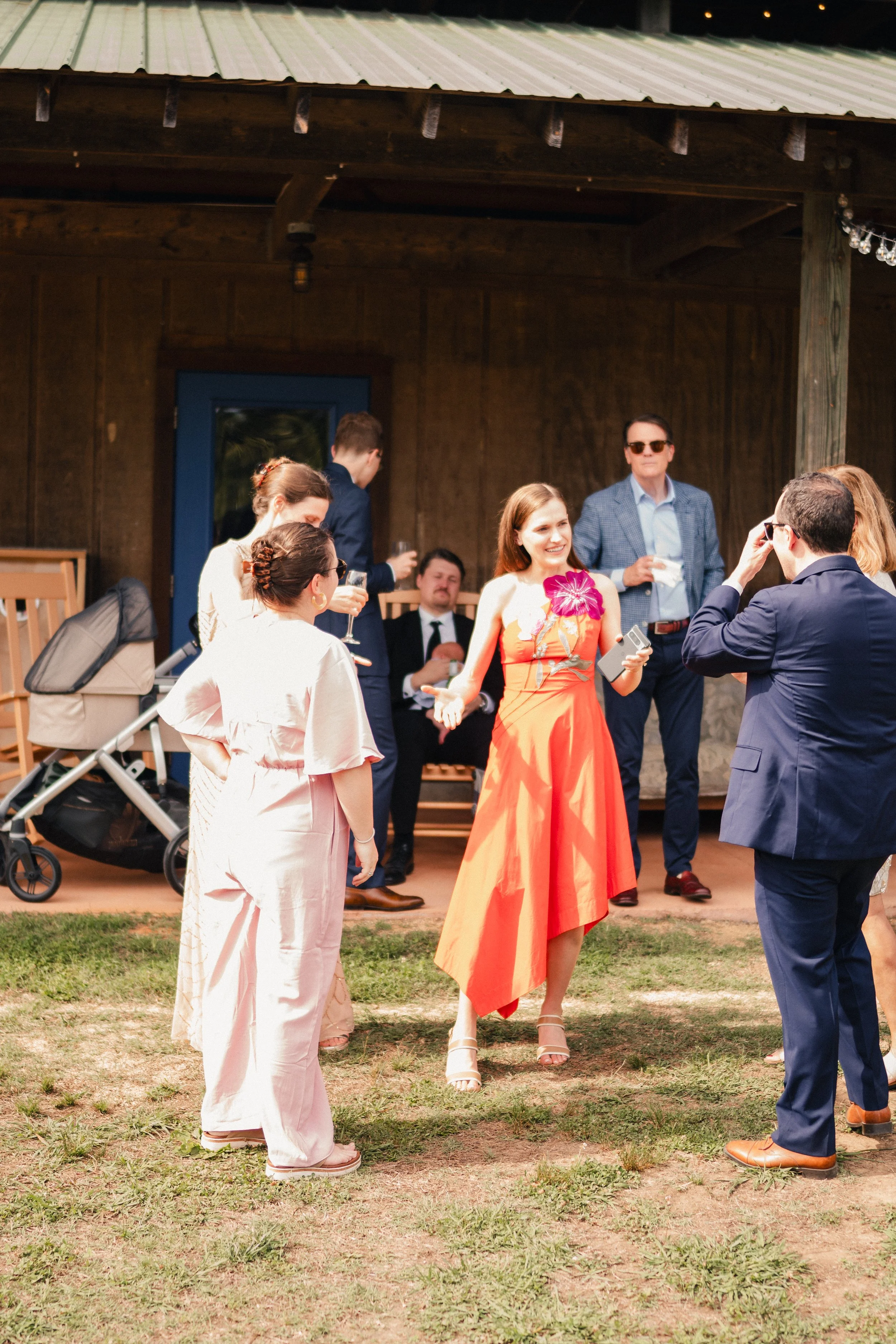 People socializing outdoors at a daytime event, with some holding drinks, in front of a rustic wooden building.