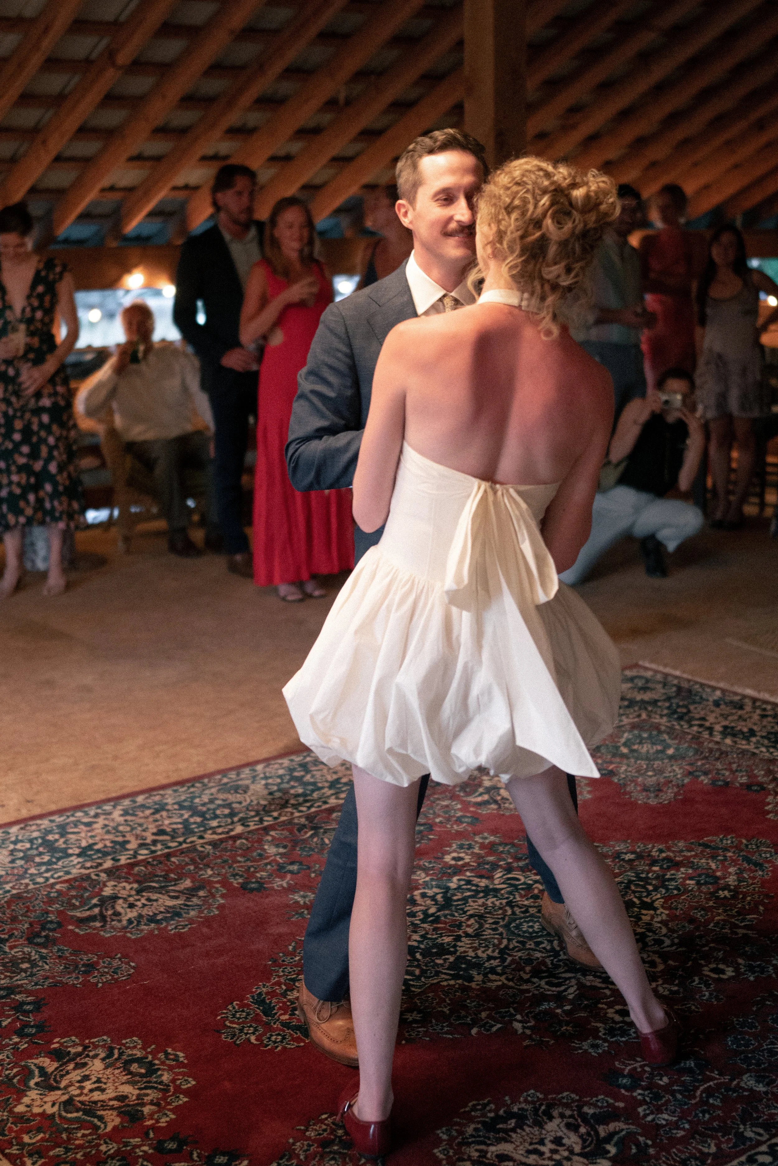 A couple dancing closely at a wedding reception, with the woman wearing a strapless white dress and red shoes, and the man in a dark suit. Several guests are watching in the background inside a rustic venue with wooden ceiling beams.