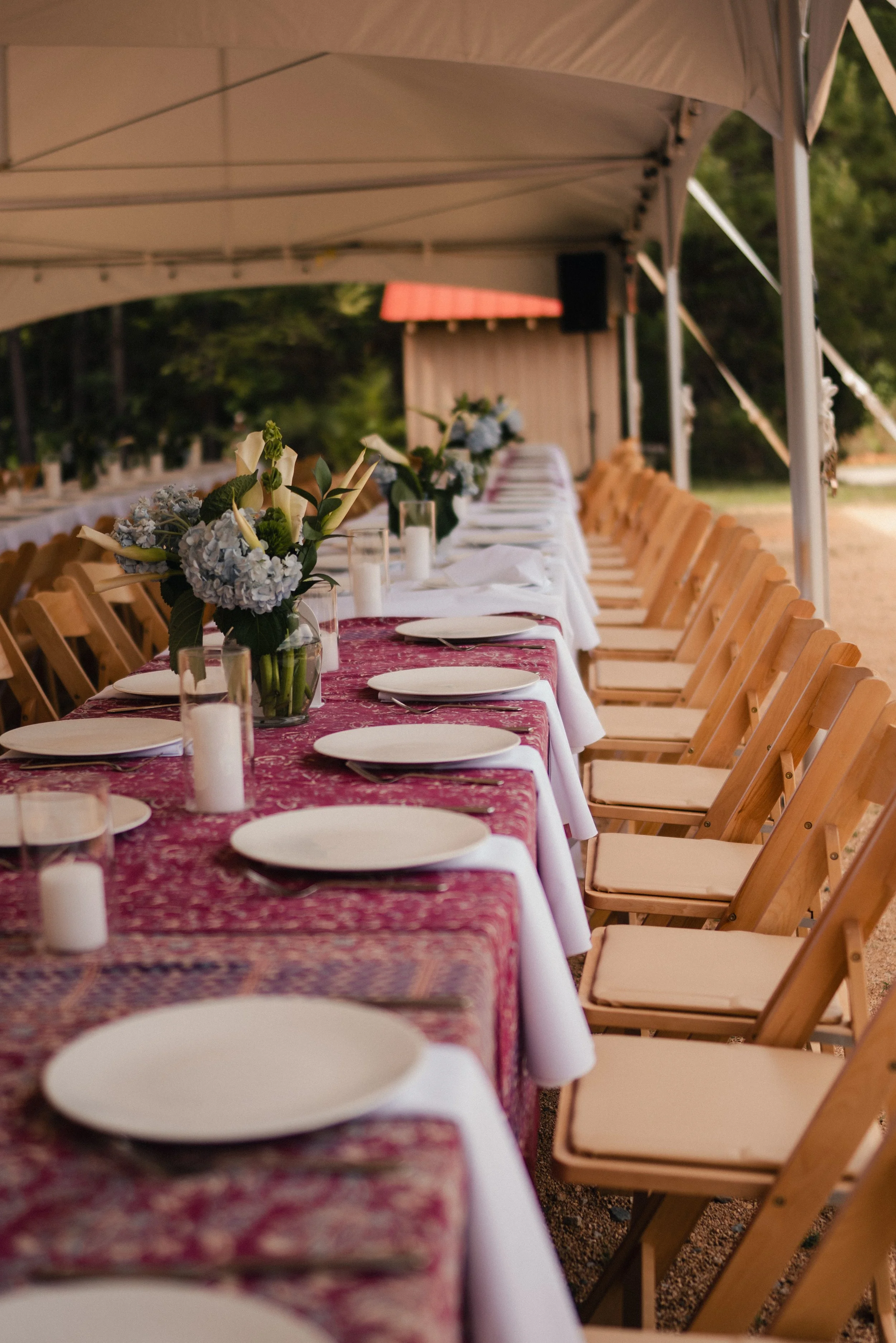 Long outdoor dinner table set under a white canopy with floral centerpieces and candles, surrounded by wooden chairs, in a garden setting.