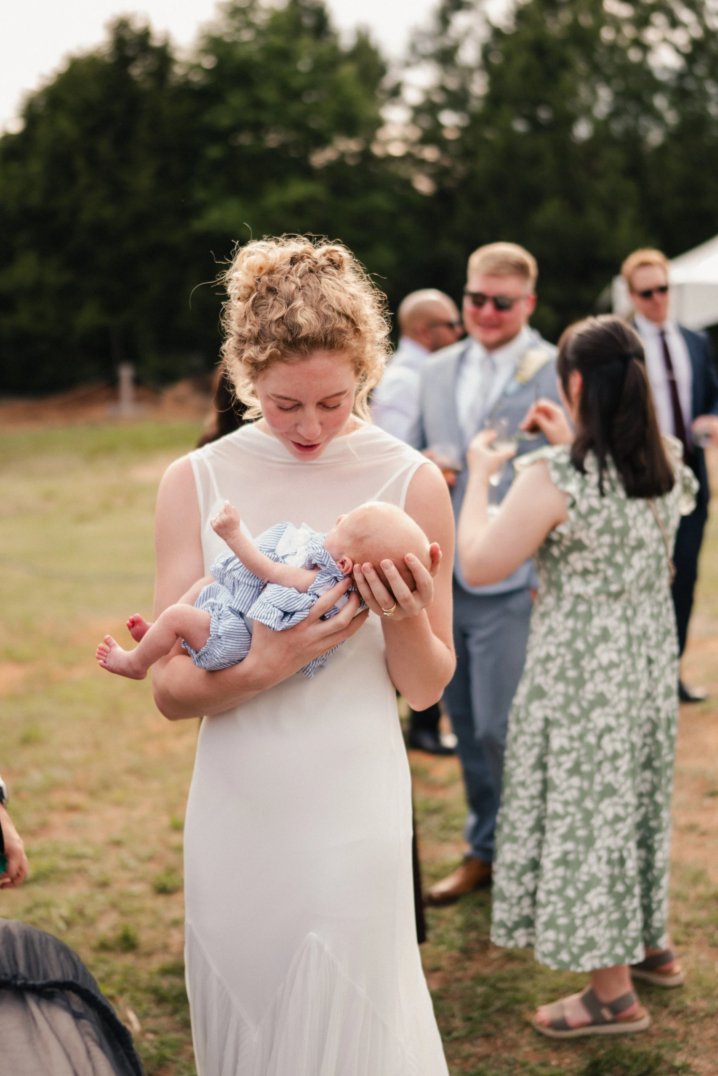 A woman in a white dress holding a baby with a striped outfit at an outdoor gathering. In the background, people are chatting and smiling, with trees and a tent visible.