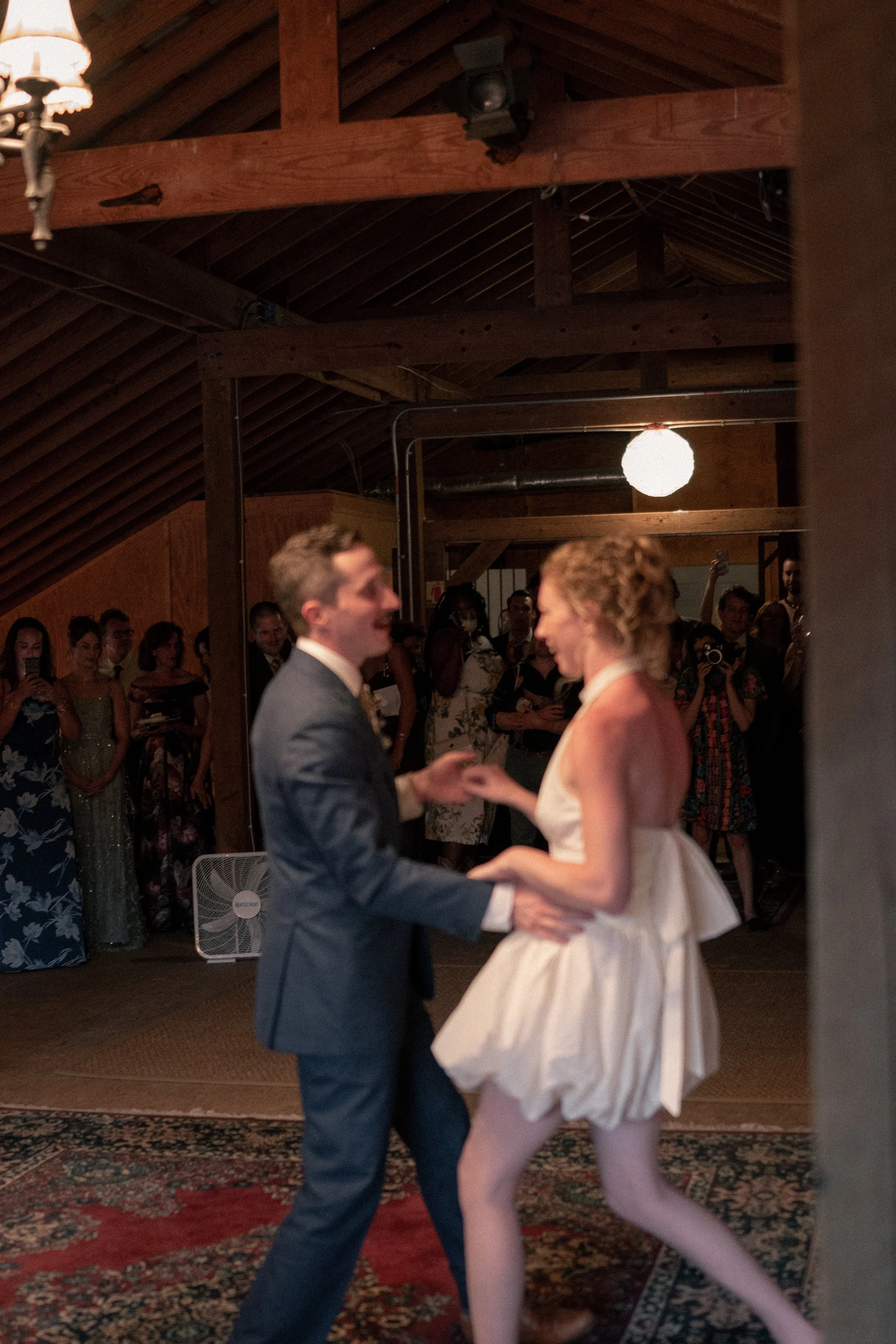 A couple dancing at a wedding reception with guests watching in the background, inside a wooden venue with a rustic ceiling and hanging lights.