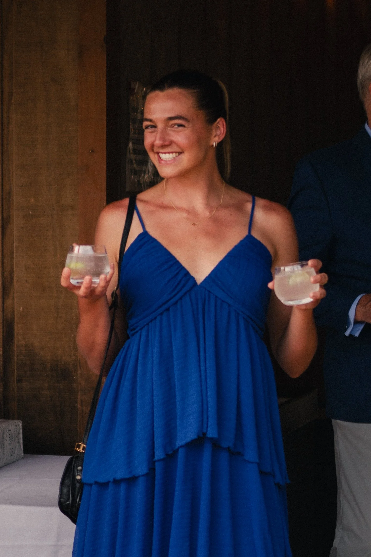 Smiling woman in a blue dress holding a clear drink in each hand at an indoor event.