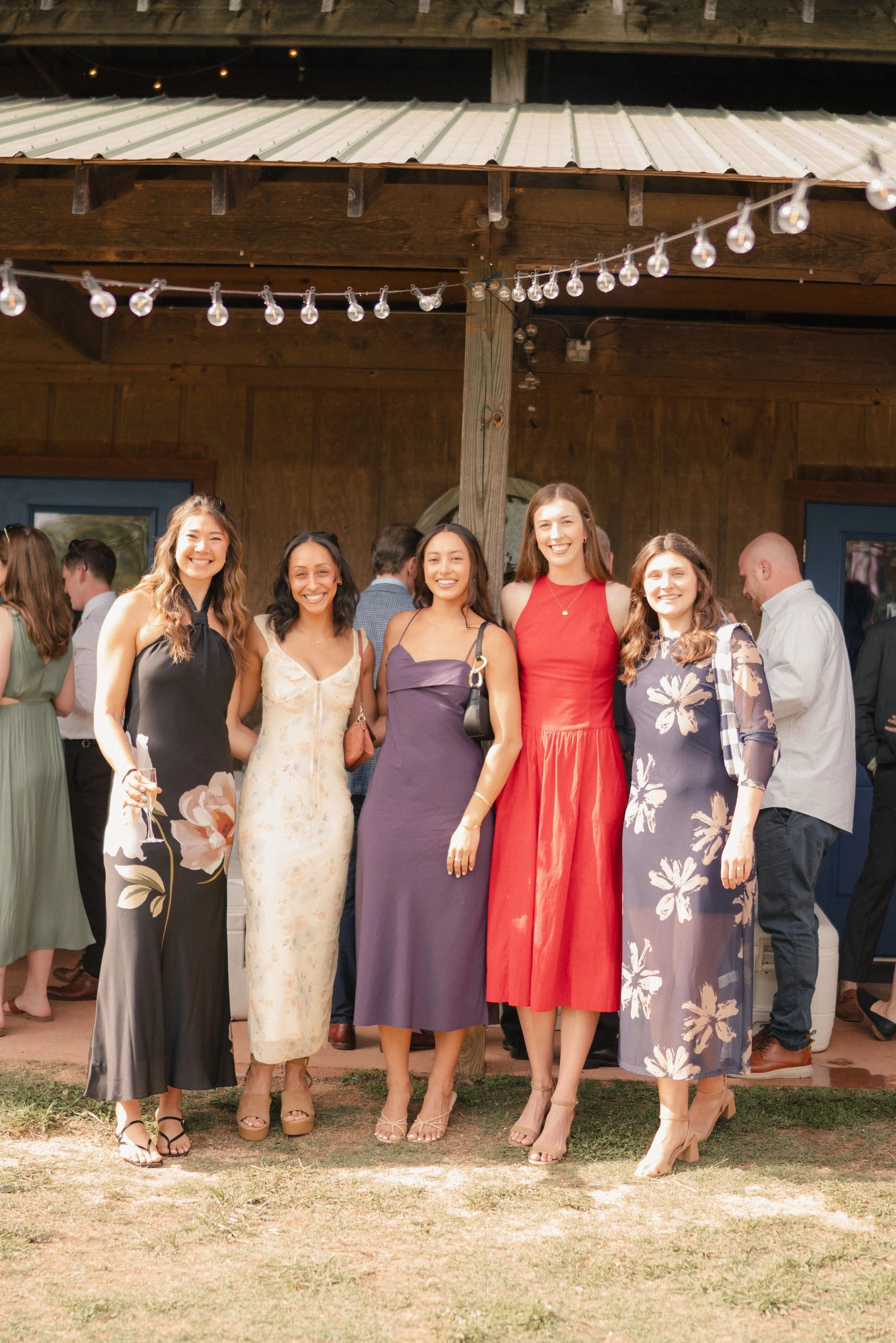 Group of five women standing outdoors at a social gathering, smiling at the camera. They are dressed in colorful dresses, and there are string lights above them, with more people and a wooden structure in the background.