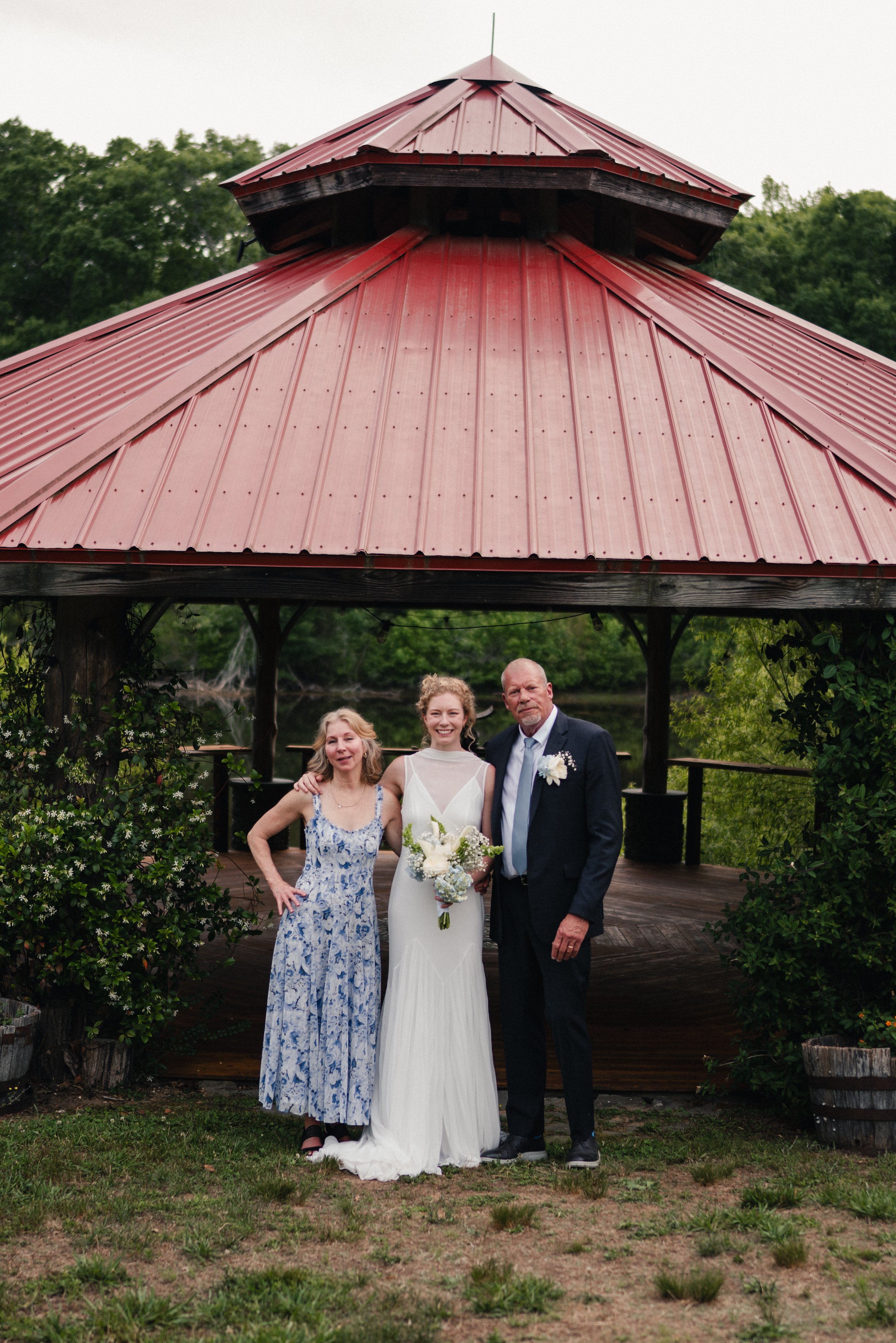 Three people standing under a gazebo with a red metal roof. Two women and a man, dressed in formal attire, are smiling for the photo. The woman in the center is wearing a white wedding dress and holding a bouquet of flowers. The other woman is in a b