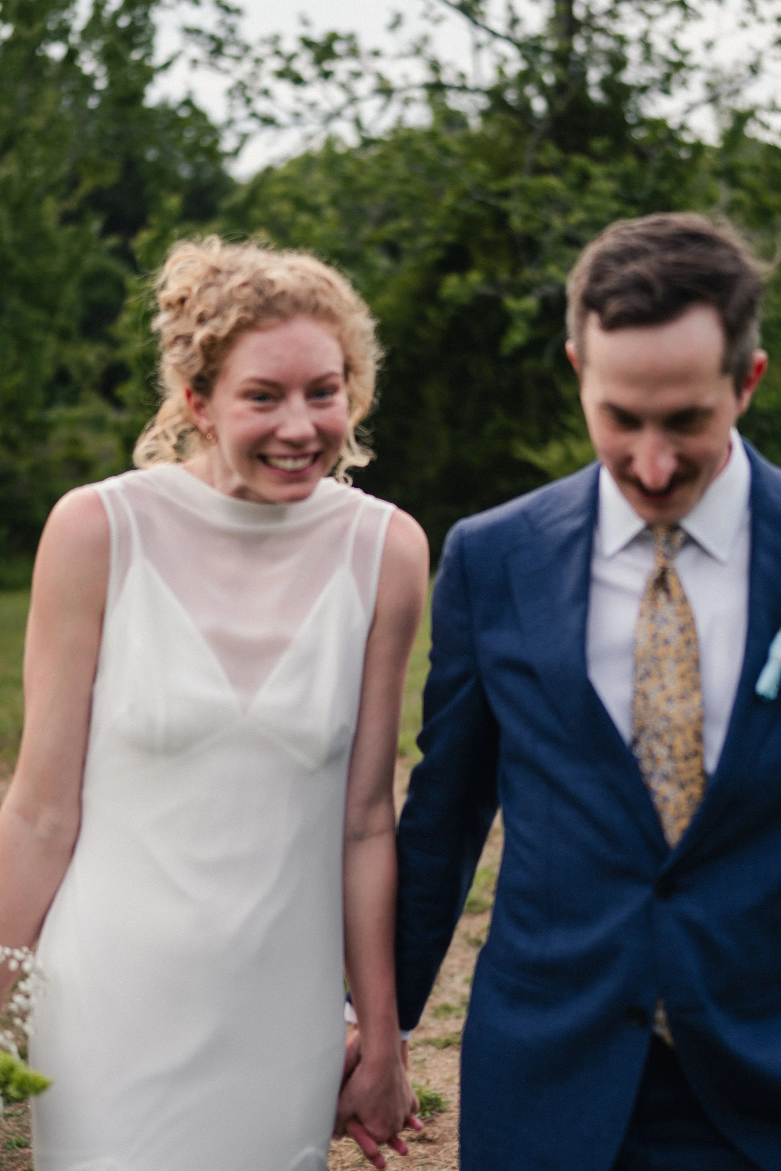 A smiling woman with curly blonde hair and a tan dress holding hands with a man in a blue suit and patterned tie, outdoors during daytime with green trees in the background.