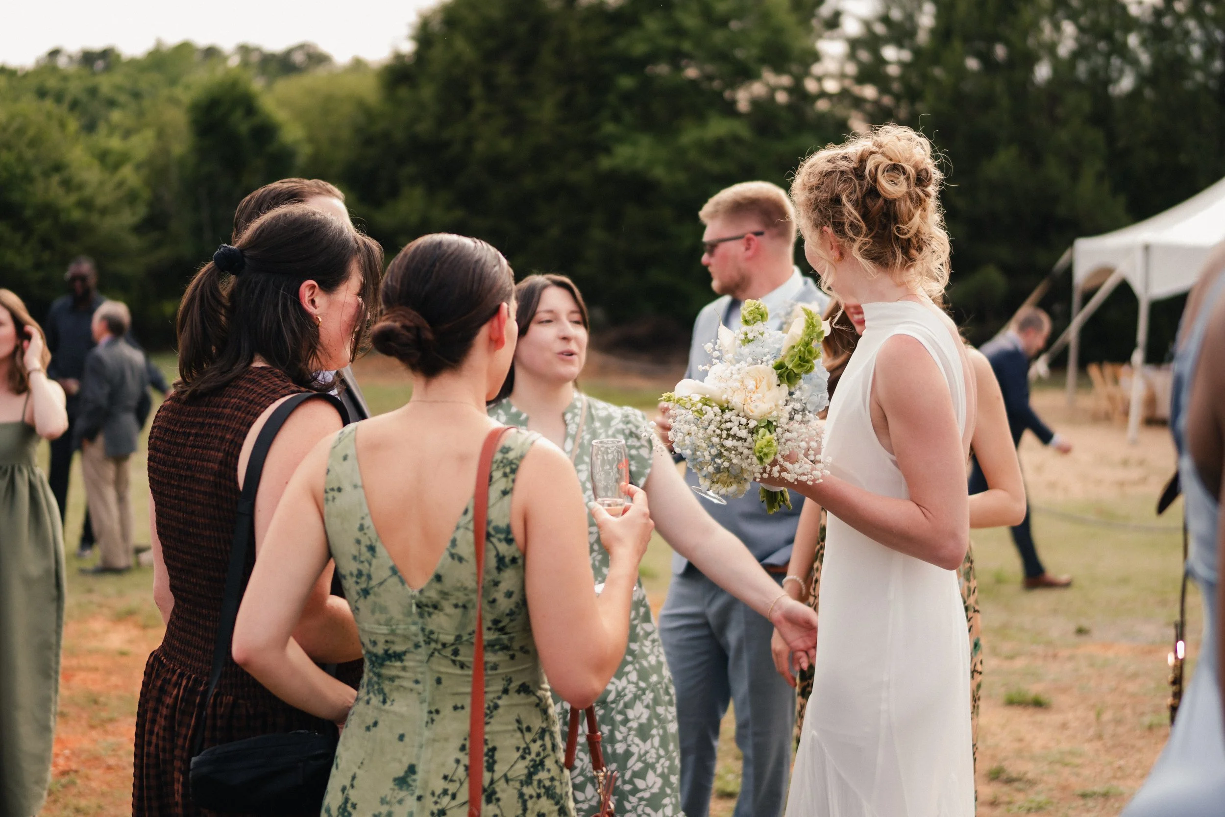 Group of people at an outdoor wedding, talking and holding drinks, with panoramic greenery in the background.