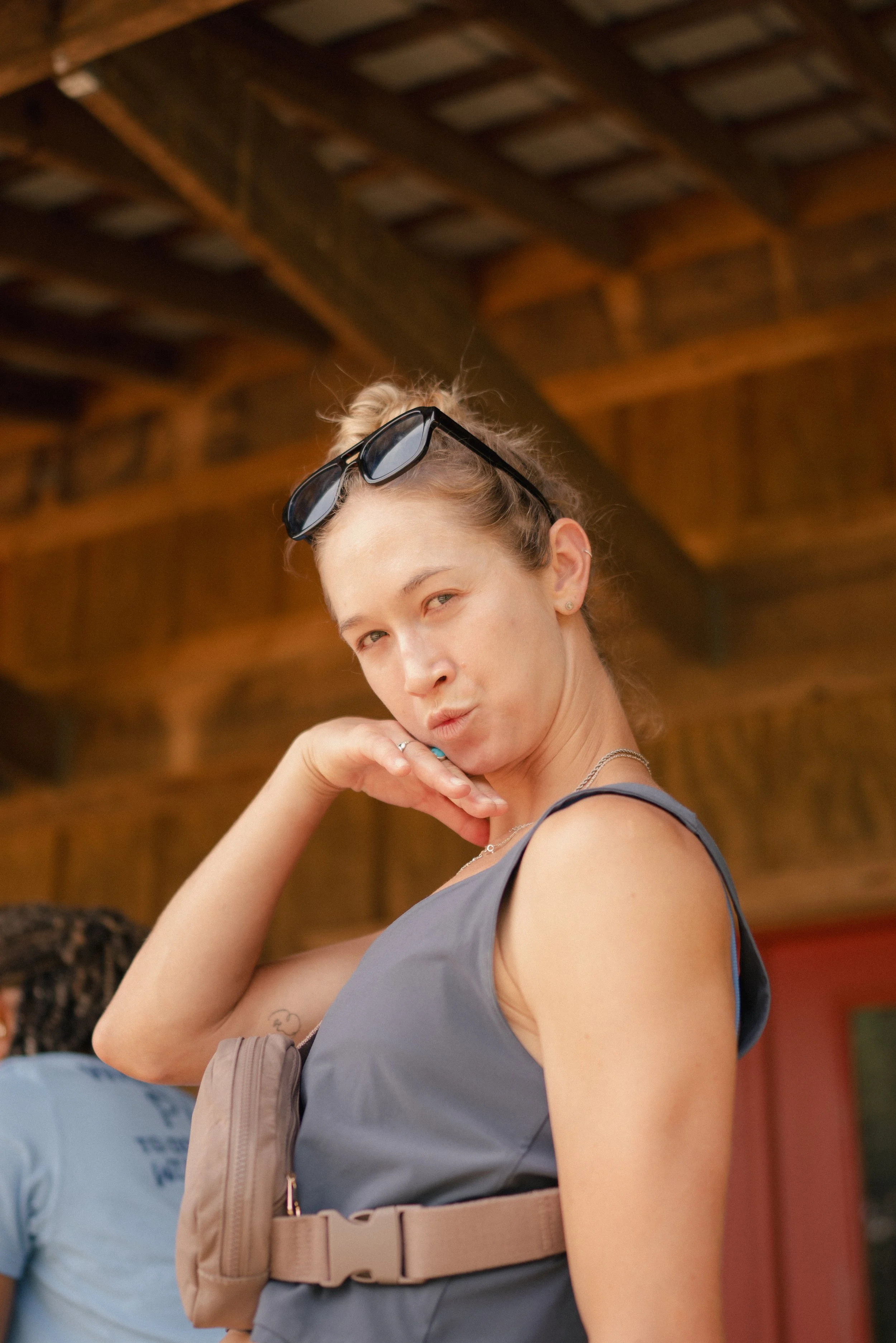 A young woman with blonde hair loose and sunglasses on her head, wearing a gray sleeveless top, standing in a wooden structure with a naturally lit background, with her hand near her face.