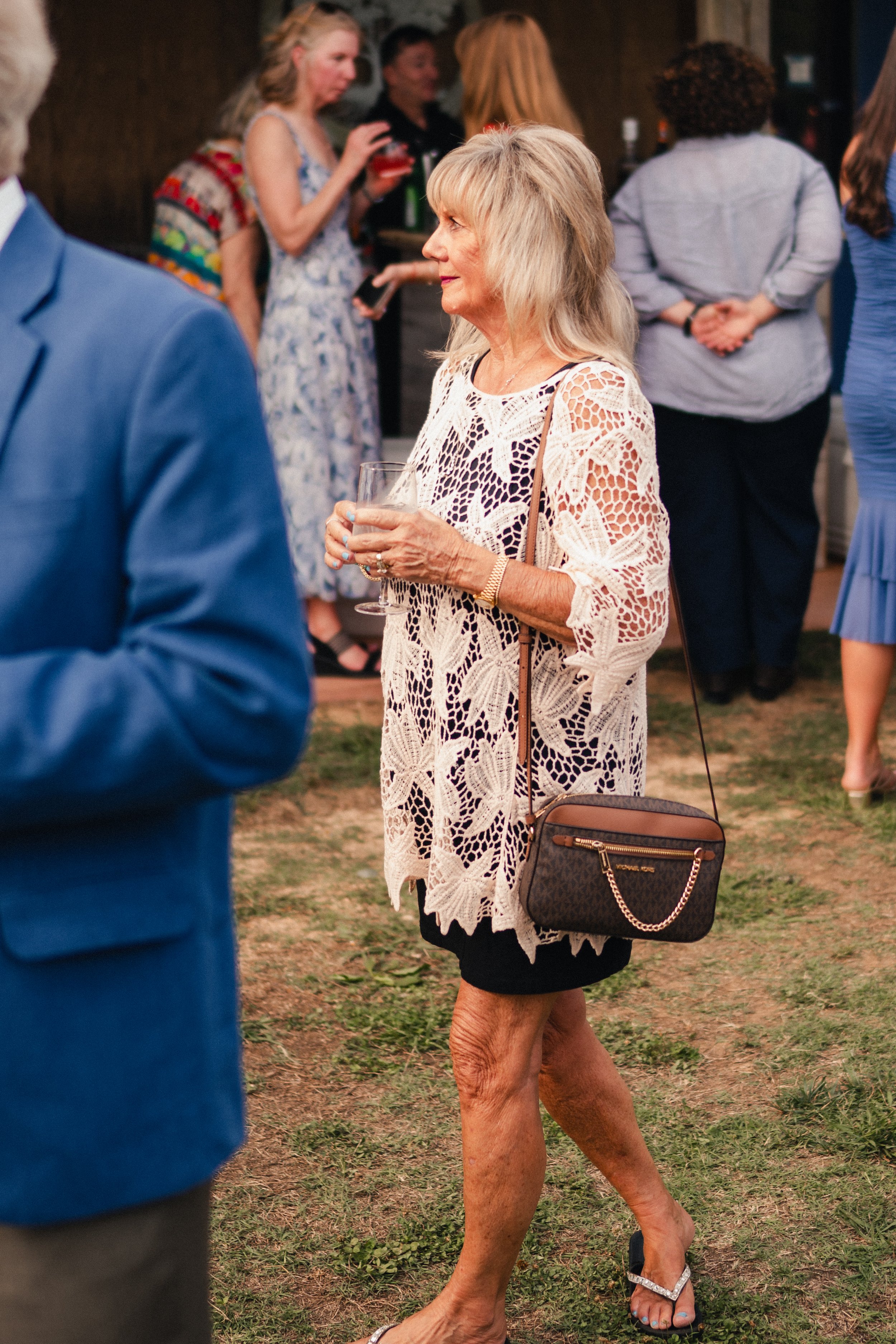 An elderly woman with long, grayish hair wearing a beige lace dress and black shorts, holding a wine glass at an outdoor social event. Several other people are in the background, engaged in conversation.