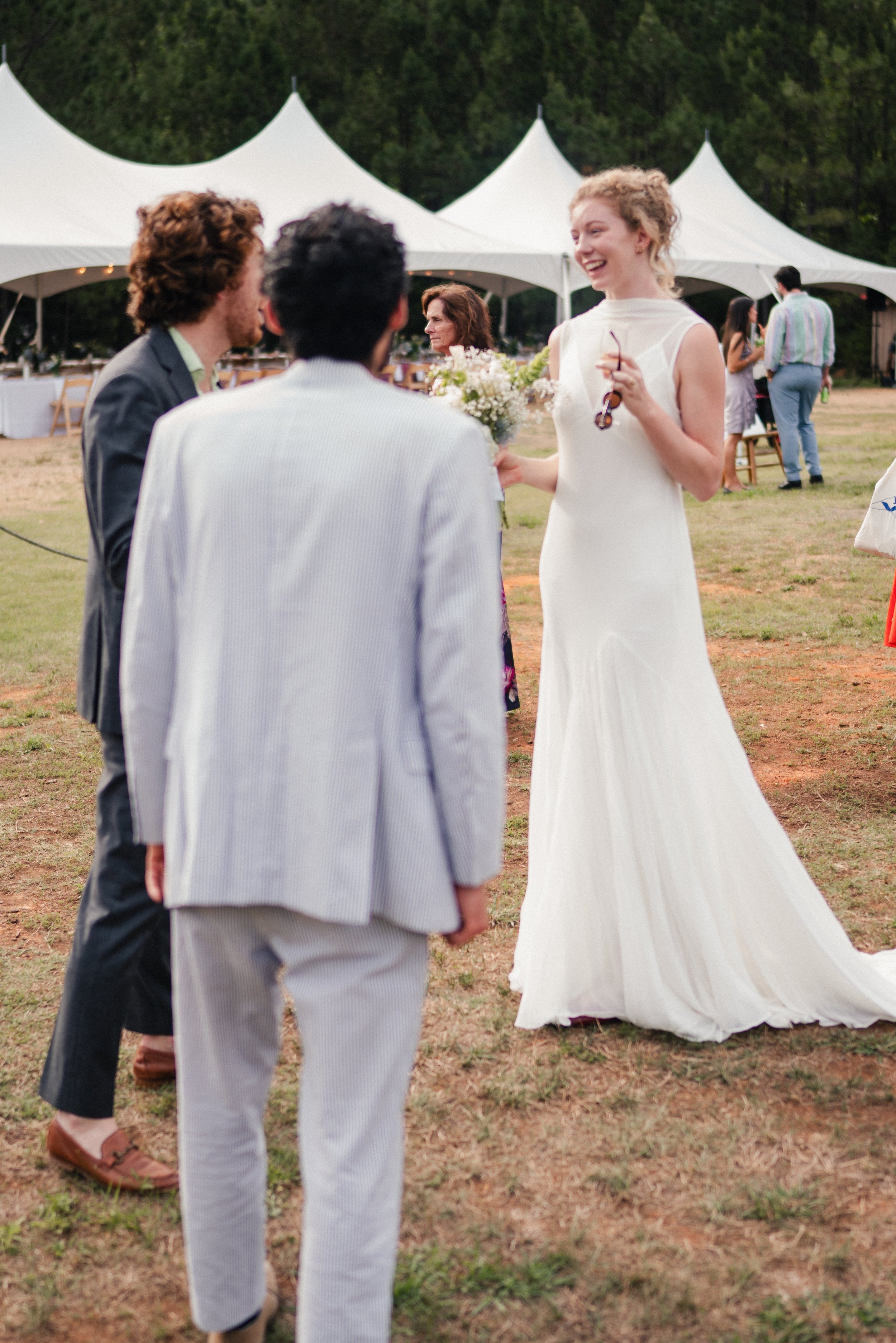 A group of people at an outdoor event, with a woman in a white dress holding flowers and smiling, talking to two men in suits. In the background, there are large white tents and other guests, with trees surrounding the area.
