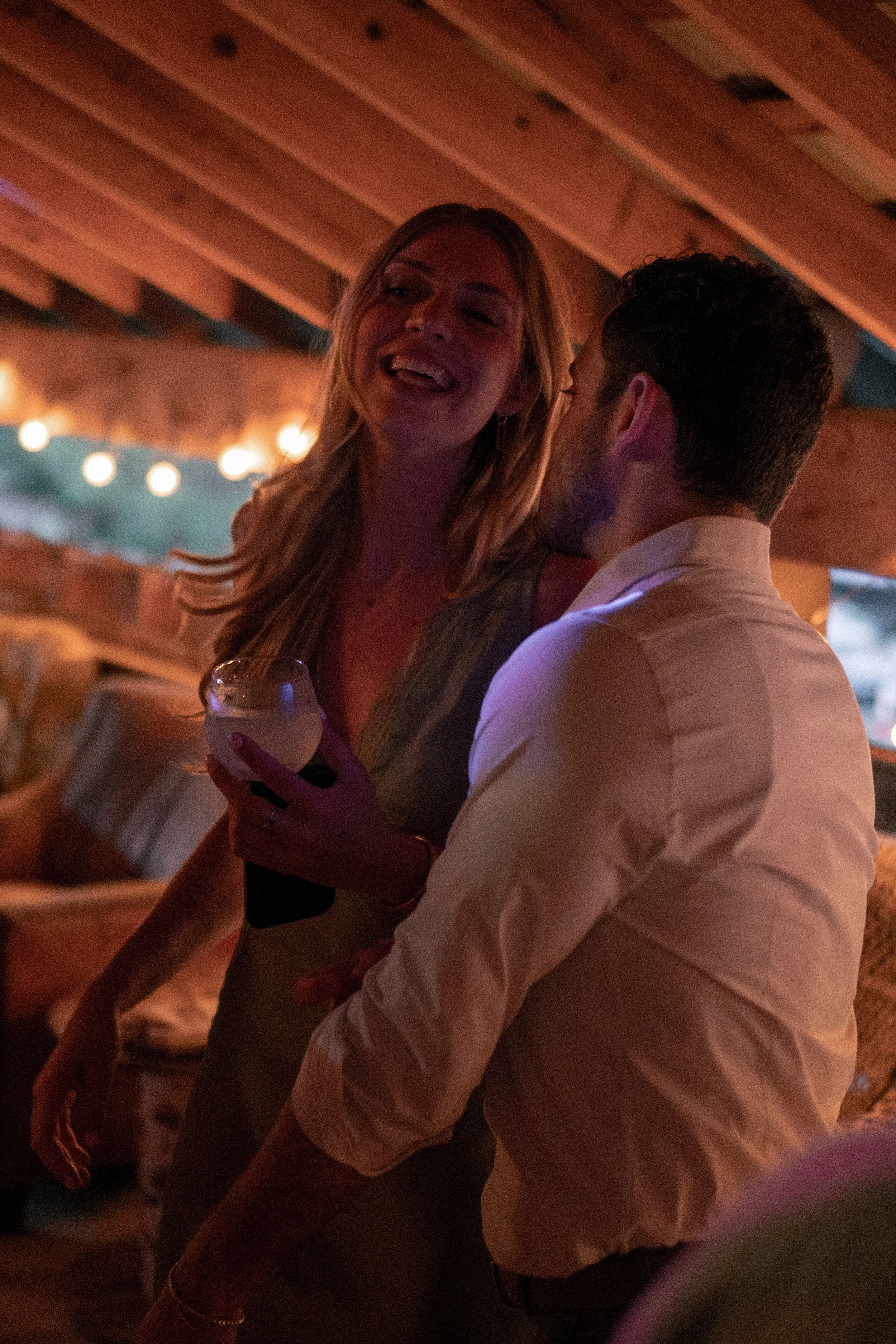 A woman with long blonde hair, smiling, is holding a glass and talking with a man with short dark hair, who is facing away. They are indoors under warm lighting, possibly at a social gathering or bar.