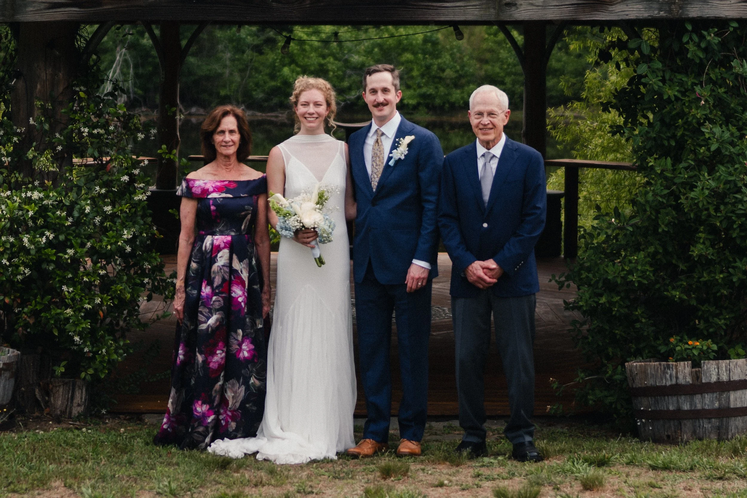 A group of five people, including a bride and groom, standing outdoors under a wooden gazebo surrounded by greenery and flowers, celebrating a wedding.