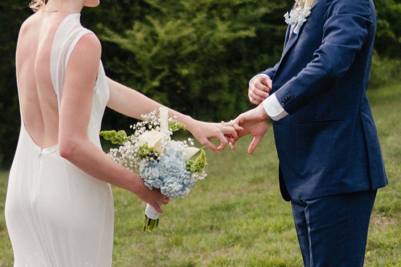 A bride and groom holding hands during a wedding ceremony outdoors, with the bride in a white dress holding a bouquet of flowers and the groom in a navy suit.