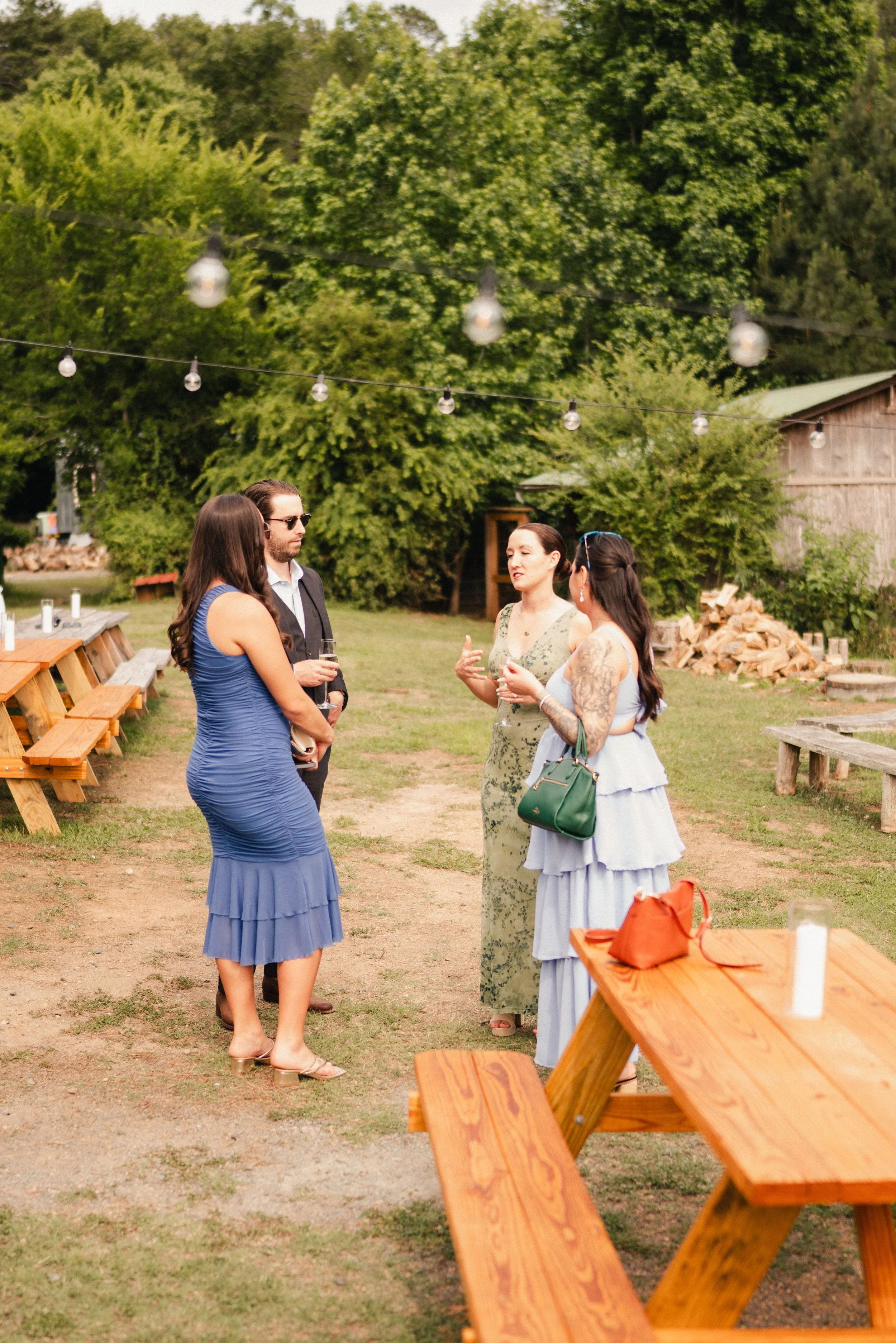 Group of four women having a conversation at an outdoor gathering, with string lights overhead and trees in the background.