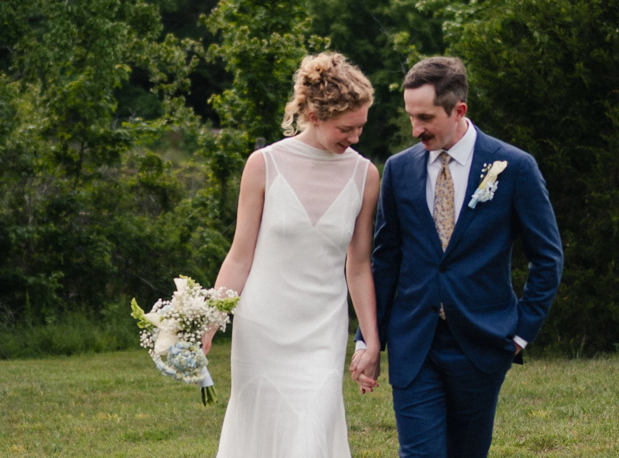 A bride and groom holding hands outdoors during a wedding ceremony, with green trees in the background. The bride wears a white dress and holds a bouquet, the groom wears a blue suit with a boutonniere.