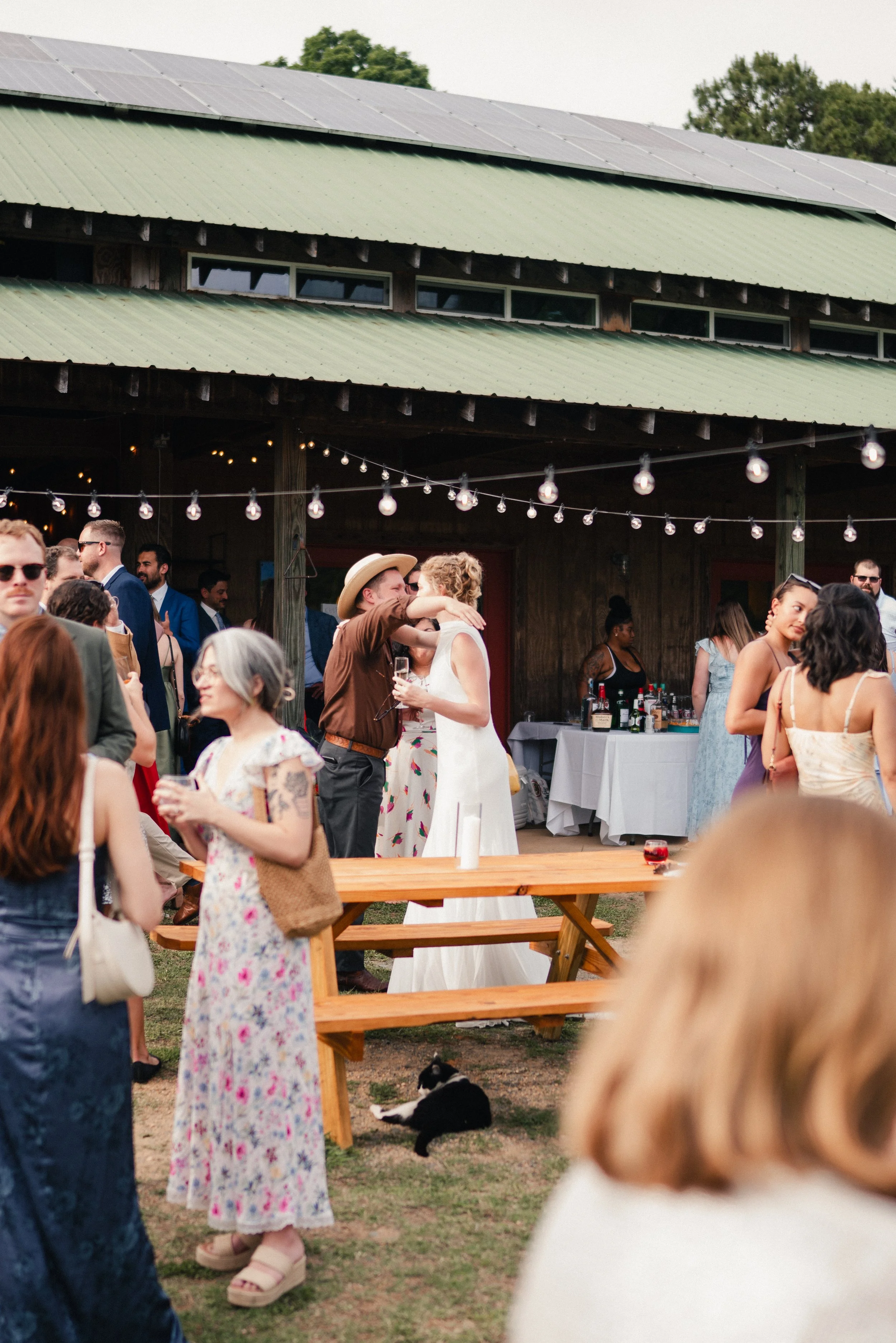 People attending outdoor wedding reception under a barn-style building with string lights, with a bride and groom dancing in the center, guests socializing, and a black and white dog lying on the grass.