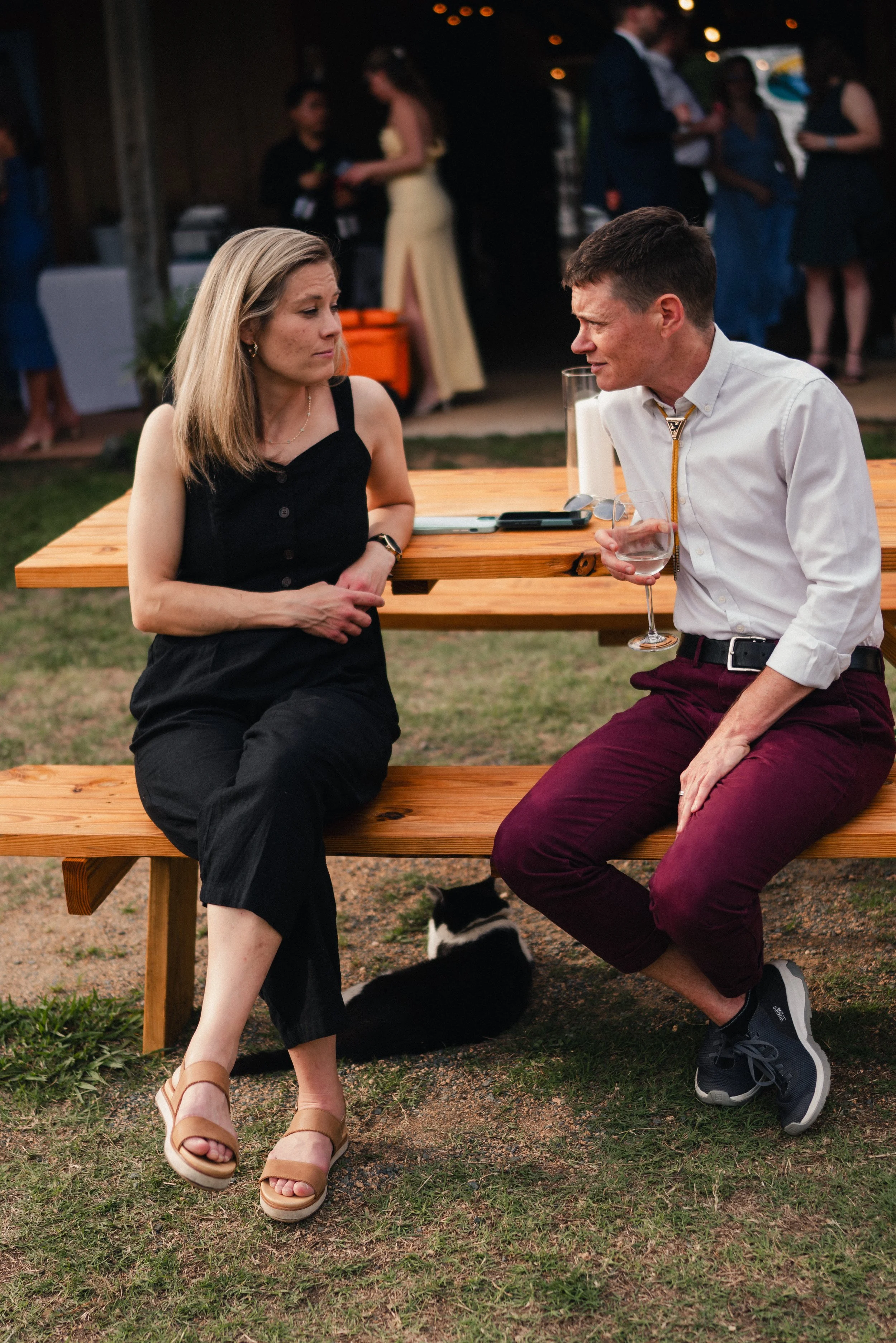 A man and woman are sitting at a wooden picnic table outdoors, engaged in conversation. The woman is wearing a black sleeveless dress and beige sandals, while the man is dressed in a white shirt, maroon pants, and sneakers, holding a glass of water. 