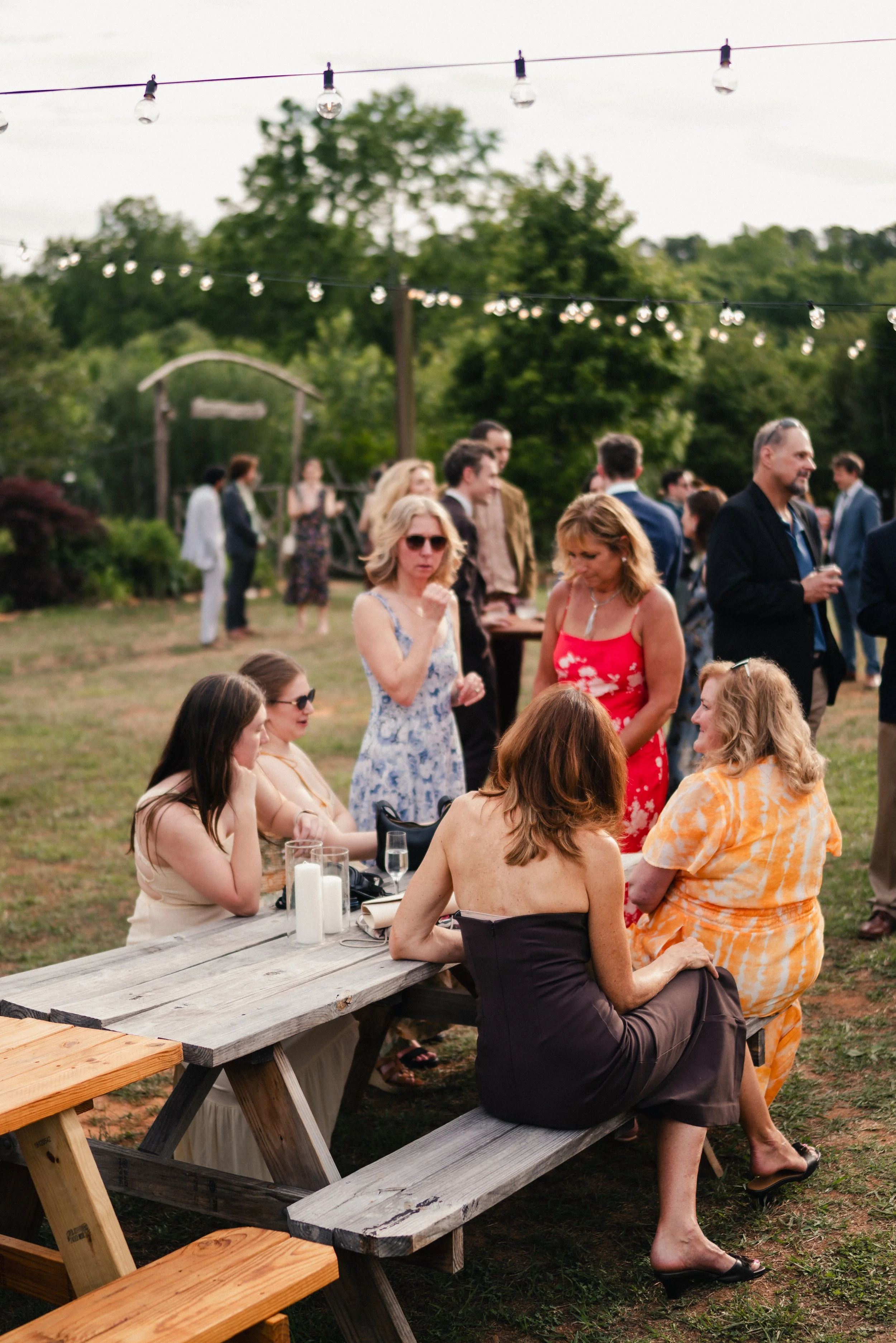 A group of people gathered outdoors during a social event, with some sitting at a wooden picnic table and others standing, chatting, and relaxing on the grass. String lights are hanging above them, and trees are visible in the background.