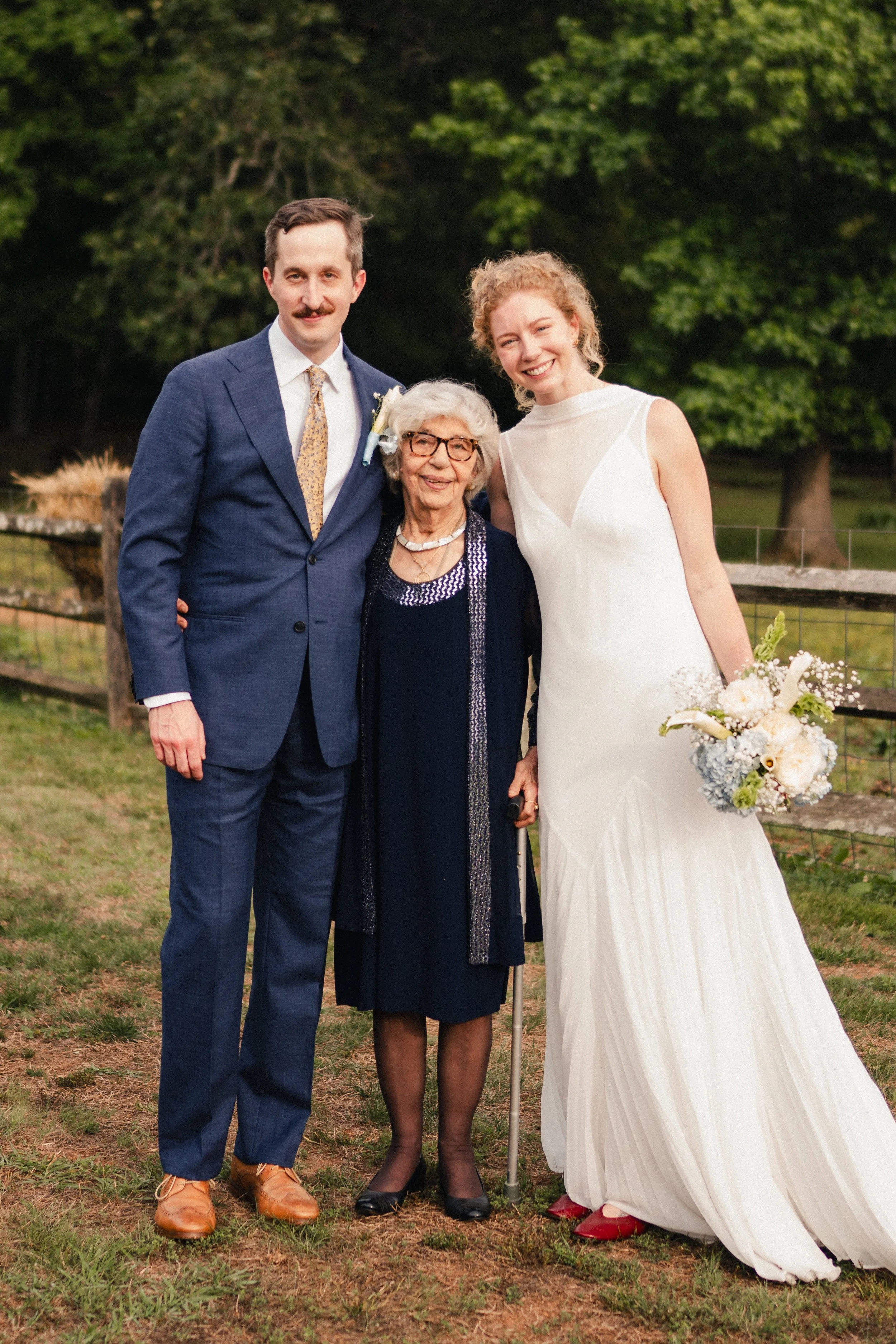 Four women standing outdoors on grass, with trees in the background. The woman on the left is wearing a navy suit with a mustard tie. The woman next to her is wearing a black dress with glasses and a white hair accessory. The third woman is wearing a