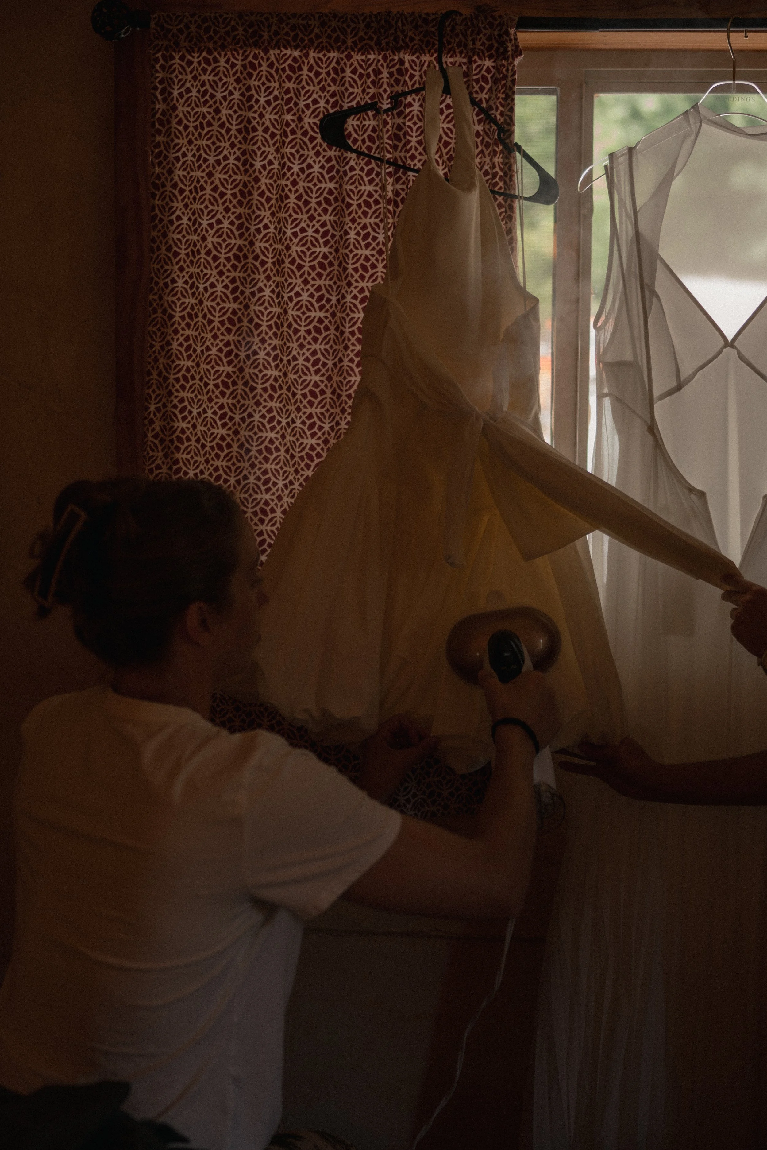 People hanging wedding dresses on a clothesline inside a room near a window with patterned curtains.