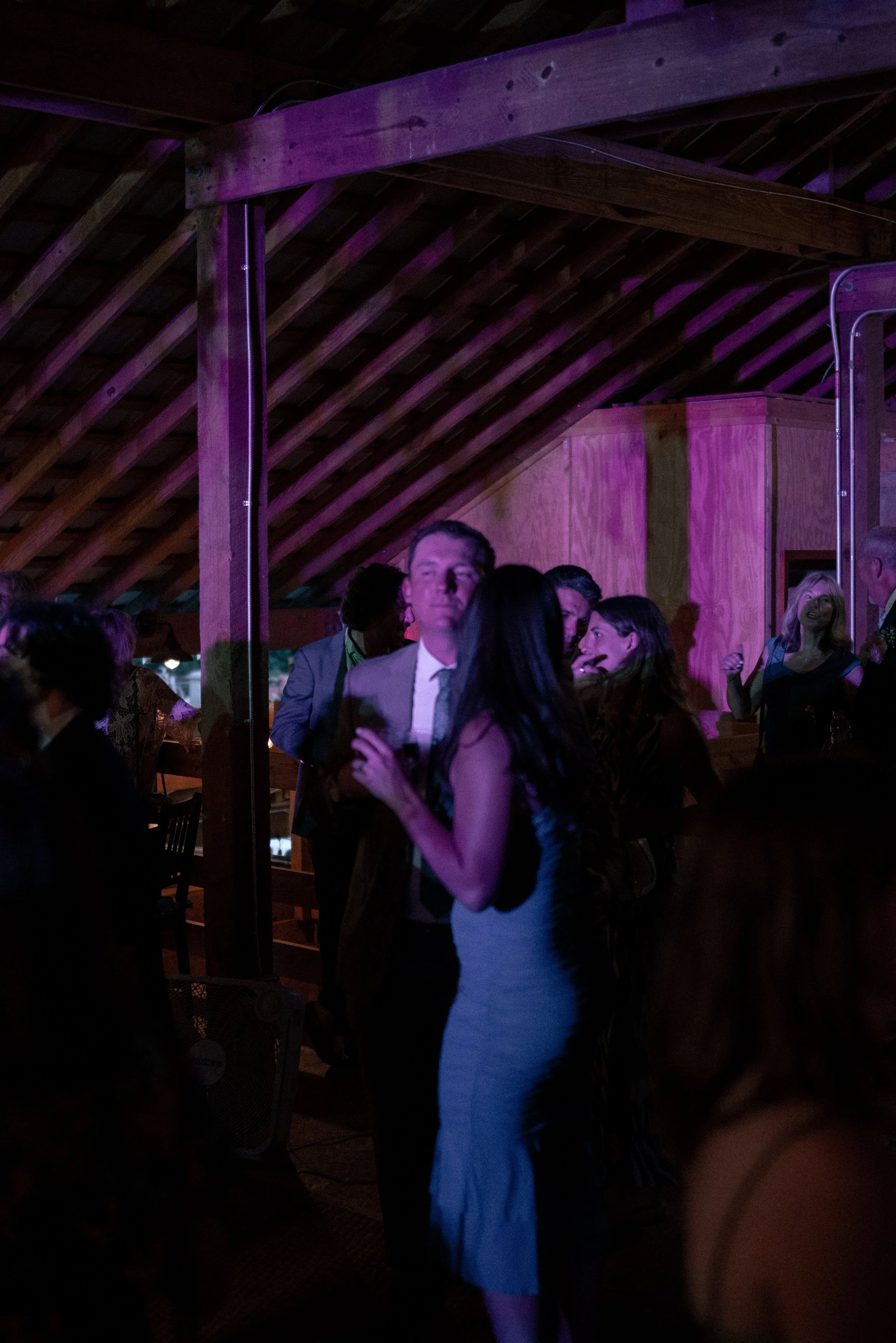 People socializing at an indoor event with wooden ceiling beams and purple lighting.