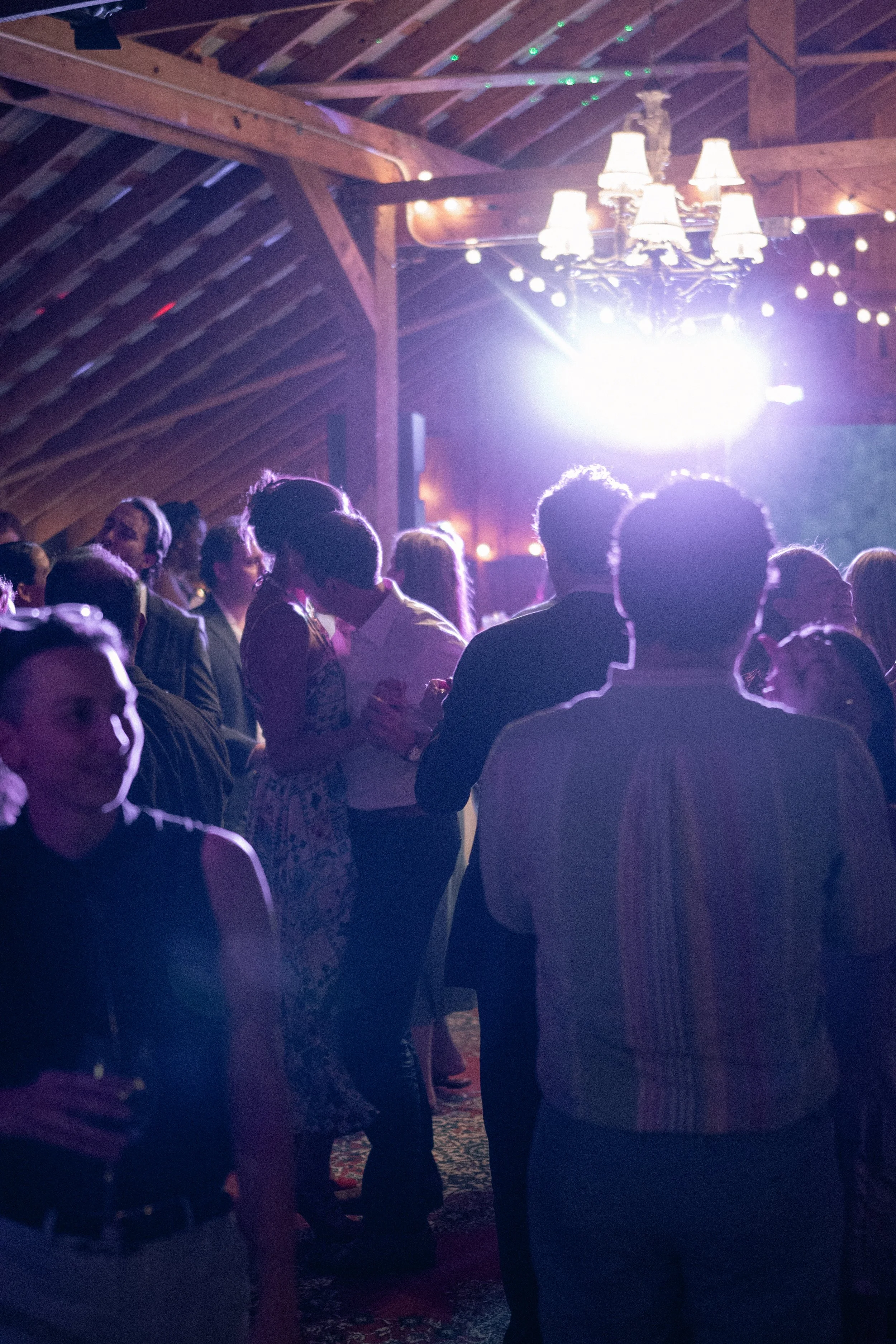 People gathering at an indoor event with wooden ceiling beams, decorated with string lights, illuminated by a chandelier, with light flare in the background.