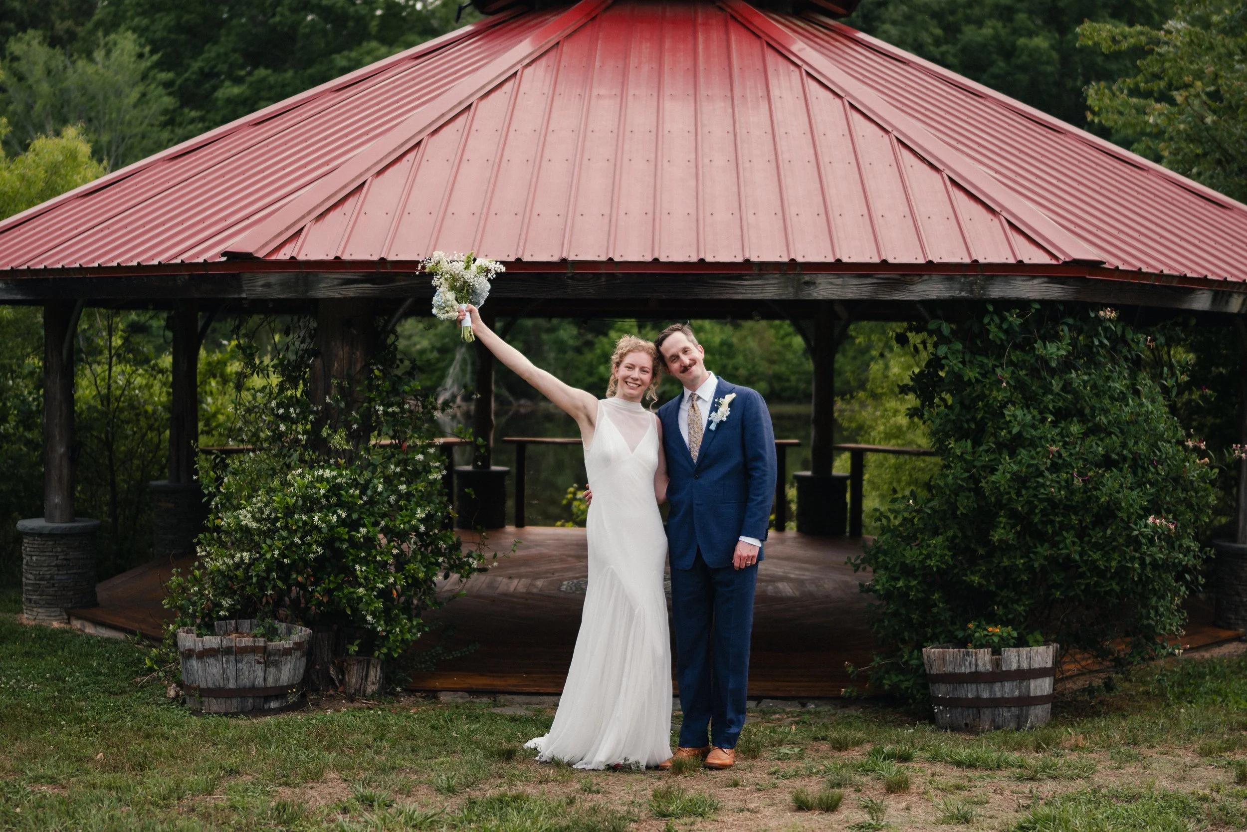A newlywed couple standing outdoors in front of a gazebo with a red metal roof. The bride in a white wedding dress is holding a bouquet of white flowers and smiling, with her arm raised in celebration. The groom in a blue suit and brown shoes is smil