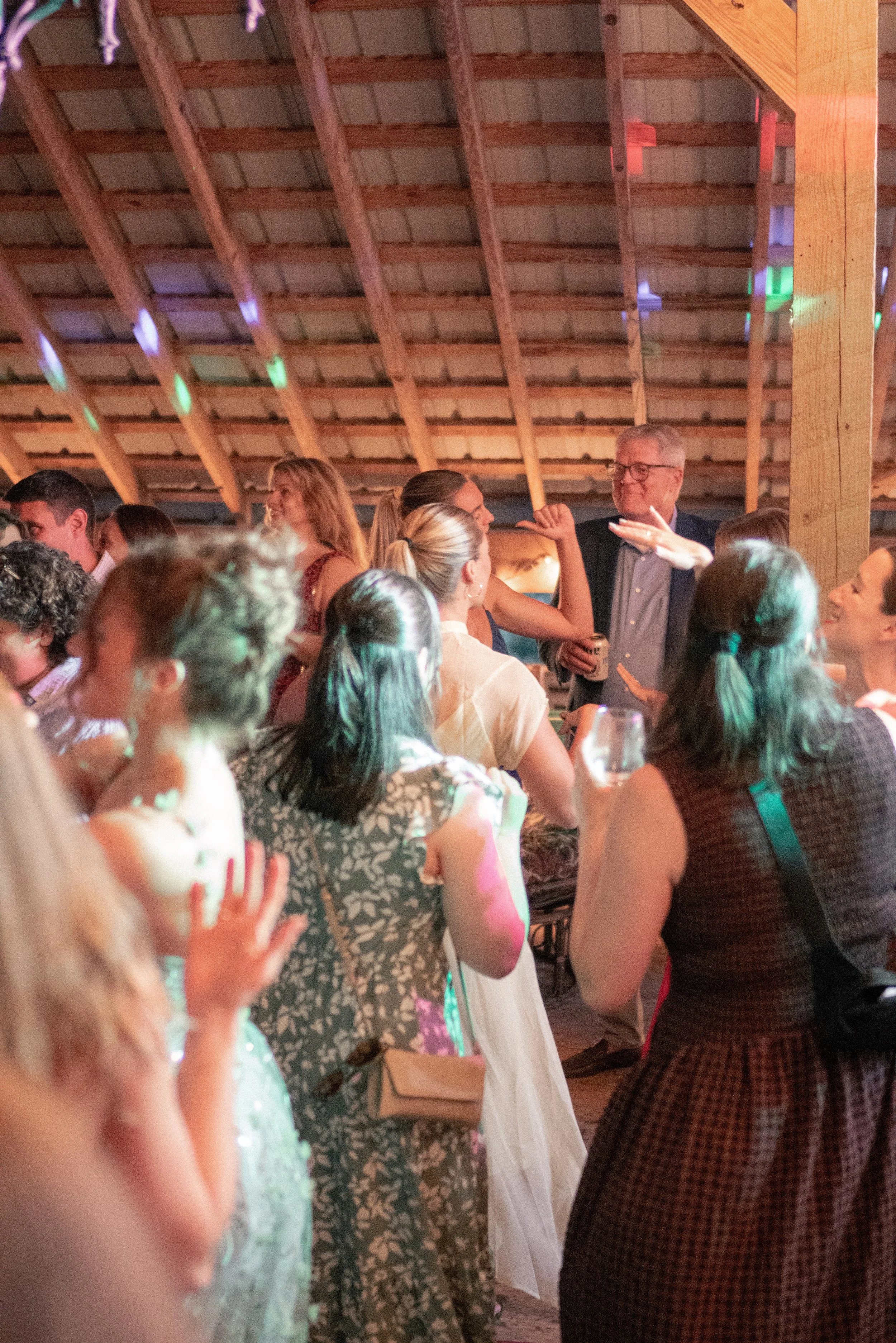 People at a social gathering or party, engaged in conversations and dancing inside a wooden venue with a sloped roof.