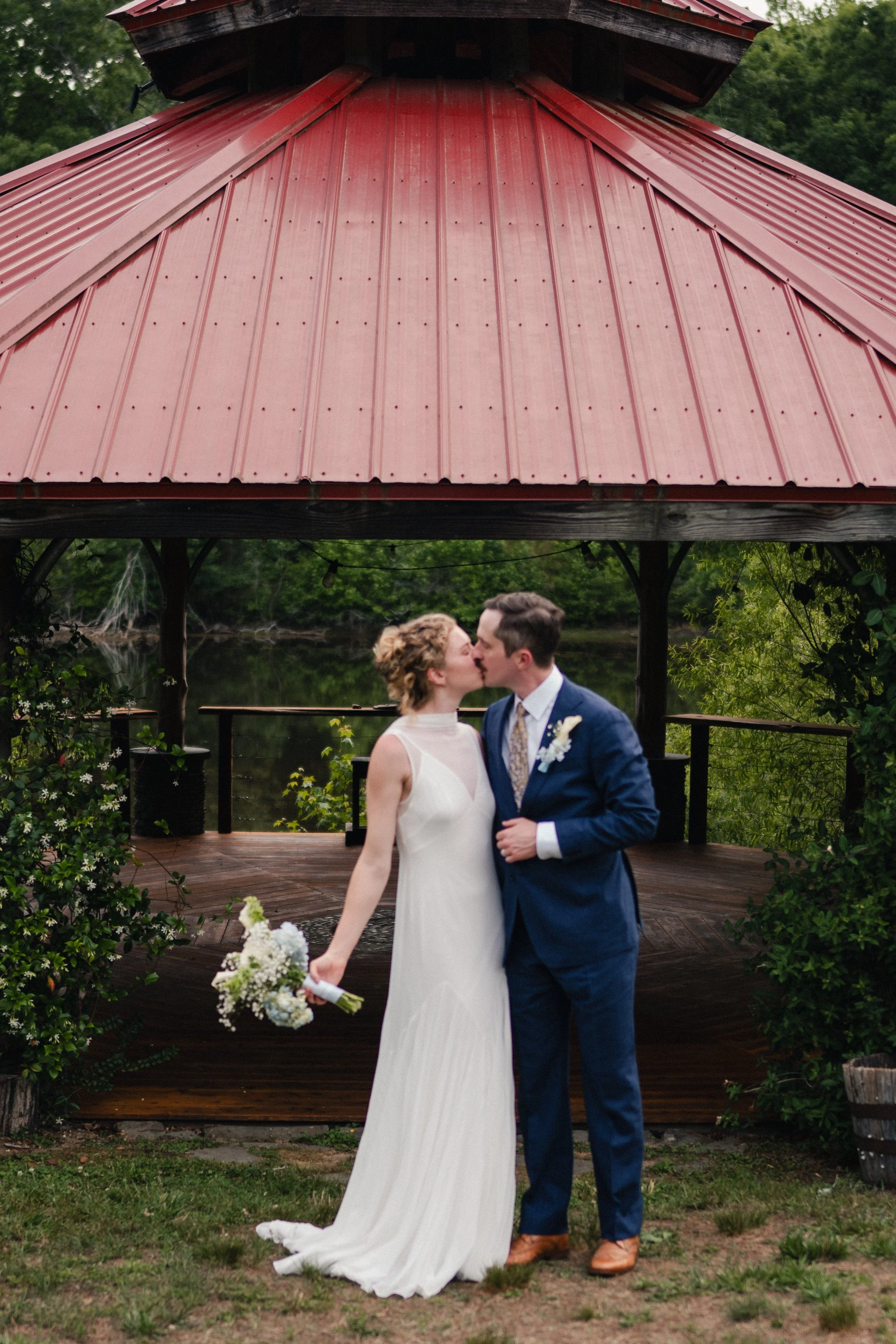 A bride and groom sharing a kiss during their wedding ceremony outdoors under a red roofed pavilion, with trees and greenery in the background.