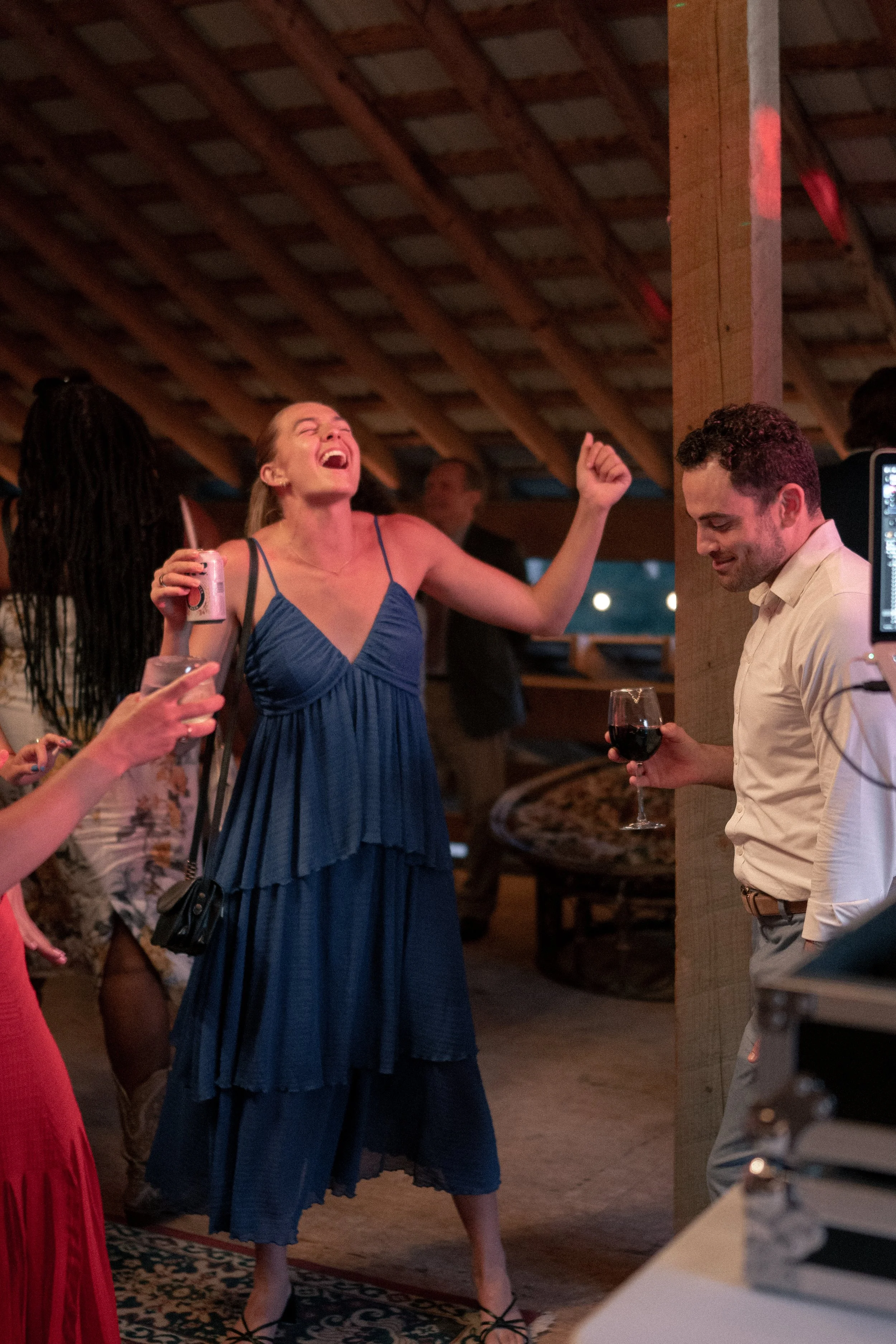 A woman in a blue dress laughing joyfully at a social gathering, holding a can of beverage, surrounded by other people in a cozy indoor setting.