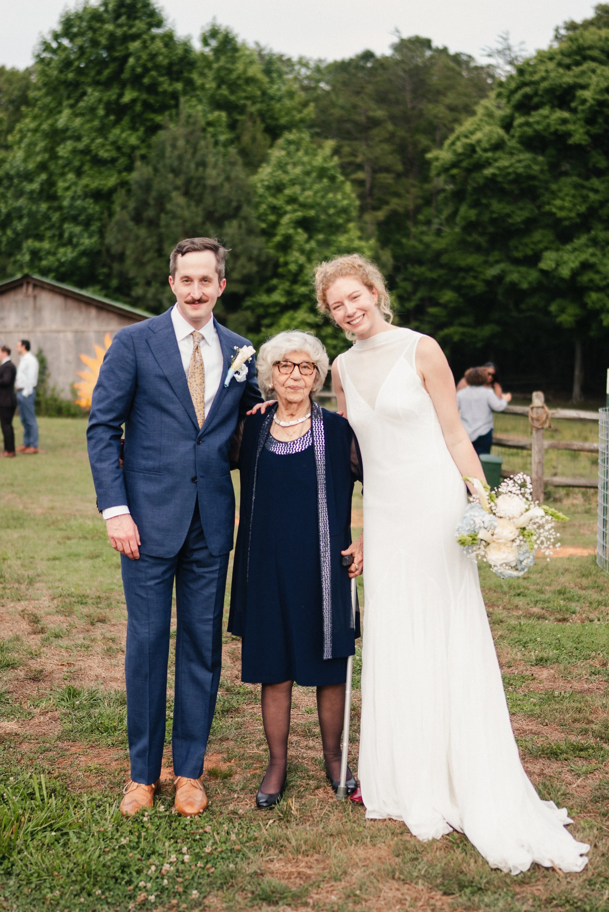 A group of four people at a wedding outdoors, with two men in suits, a woman in a black dress, and a woman in a white wedding dress holding a bouquet, standing on grass with trees in the background.